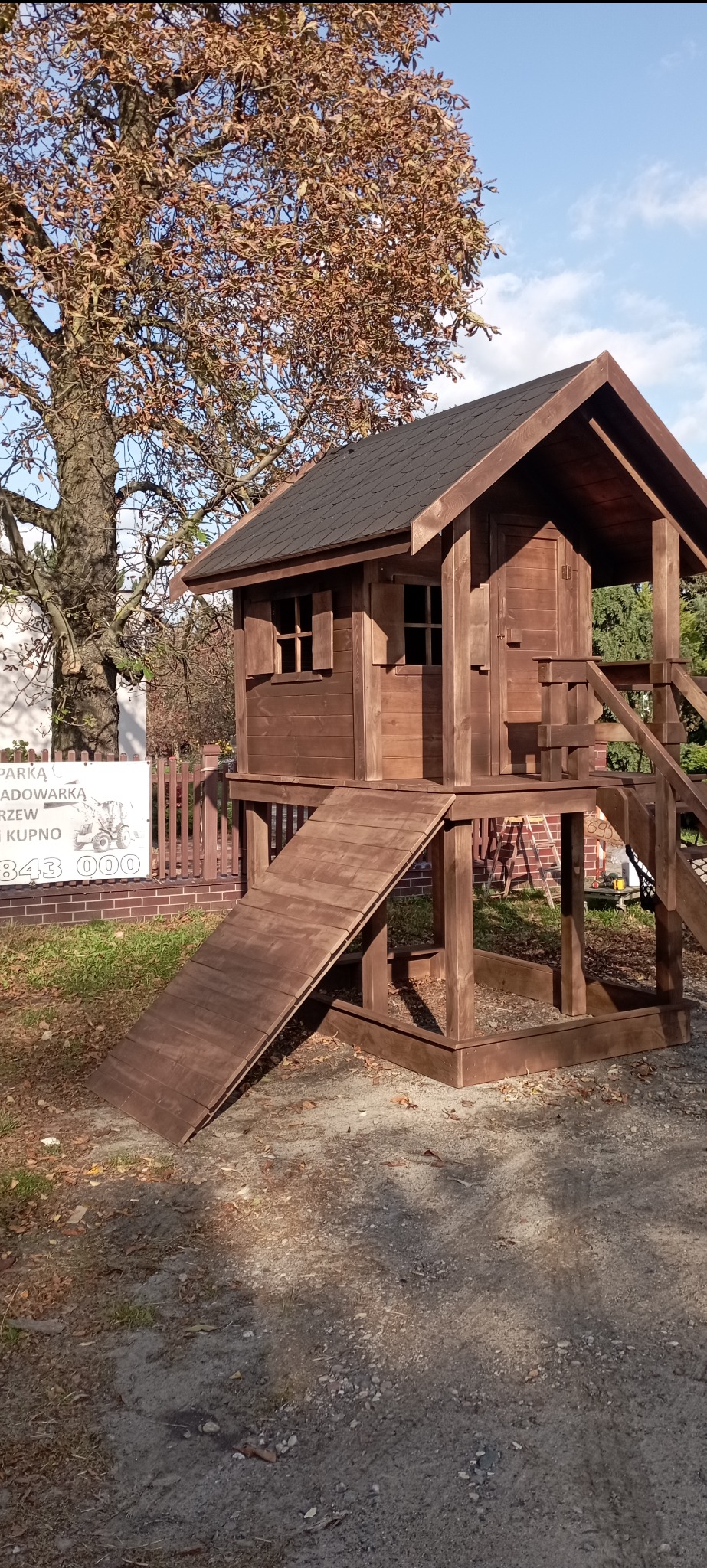 Drewniany domek dla dzieci z zadaszeniem i zjeżdżalnią, w odcieniach brązu, umieszczony na trawniku obok drzewa z brązowymi liśćmi i białej tablicy z napisem.