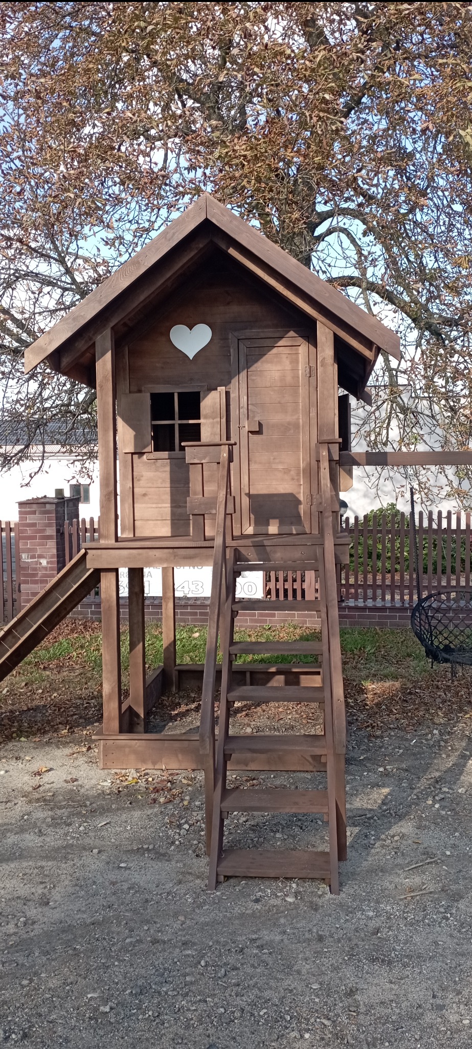 Drewniany domek zabaw dla dzieci z sercem na ścianie, schodkami i zjeżdżalnią, umieszczony na zewnątrz w ogrodzie. Widoczne drzewo w tle.
