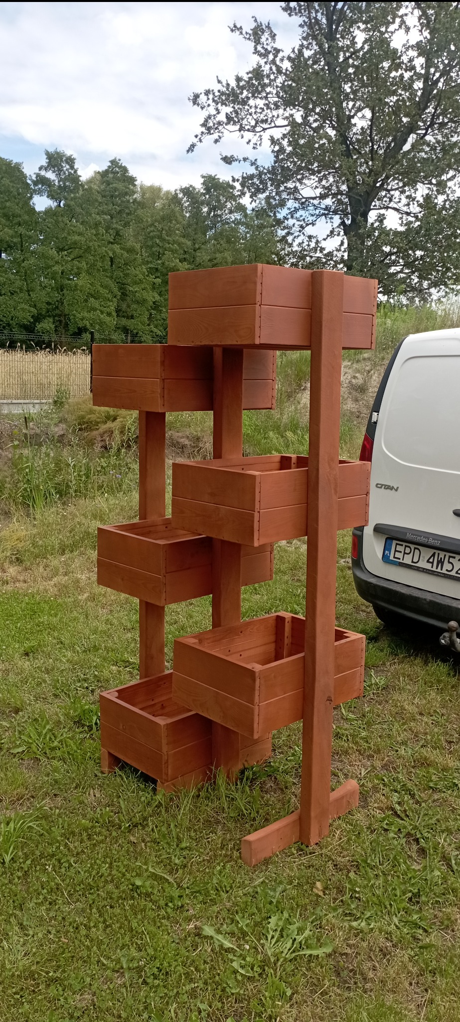 Drewniany kwietnik kaskadowy z sześcioma skrzynkami, ustawiony na trawie obok białego samochodu. Konstrukcja zaimpregnowana na brązowo, widoczne detale łączeń.