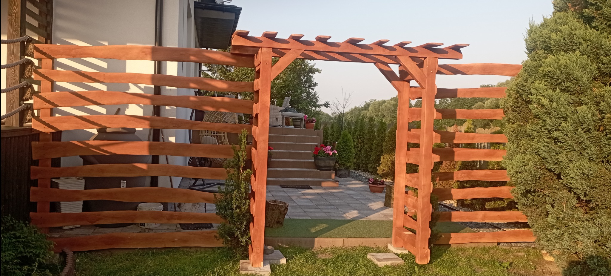 Drewniana konstrukcja z bramką i płotem z poziomych desek, prowadząca do schodów z donicami kwiatów. Elementy wykonane z drewna w ciepłym odcieniu brązu, otoczone zielenią.