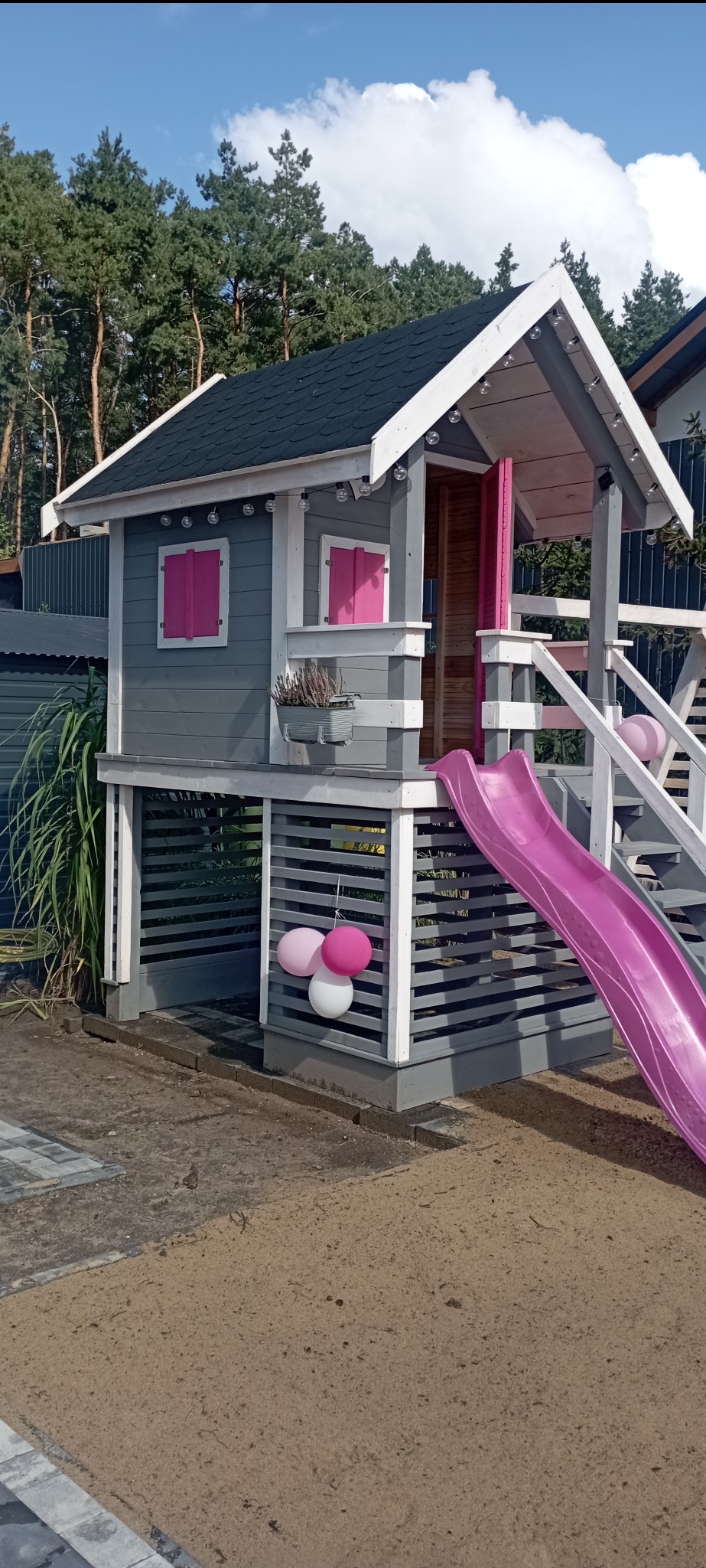 Drewniany domek dla dzieci w szaro-białej kolorystyce z różowymi akcentami, wyposażony w zjeżdżalnię i ozdobiony lampionami oraz balonami. Stoi na piaszczystym podłożu.