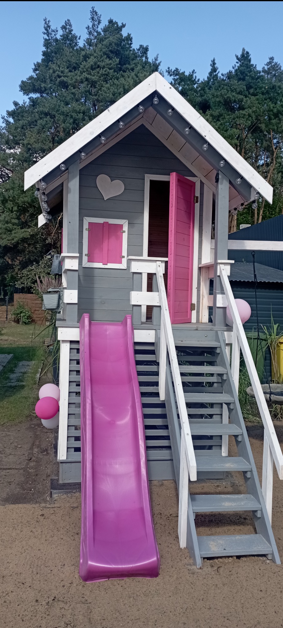 Szara altanka dla dzieci z różową zjeżdżalnią i otwieranymi drzwiami, ozdobiona sercem i lampkami, umieszczona w ogrodzie.