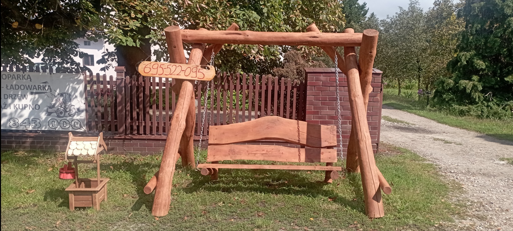 Drewniana huśtawka ogrodowa z surowego drewna, łańcuchy, tabliczka z numerem telefonu, mała studnia dekoracyjna na trawie, w tle ogrodzenie i zieleń.