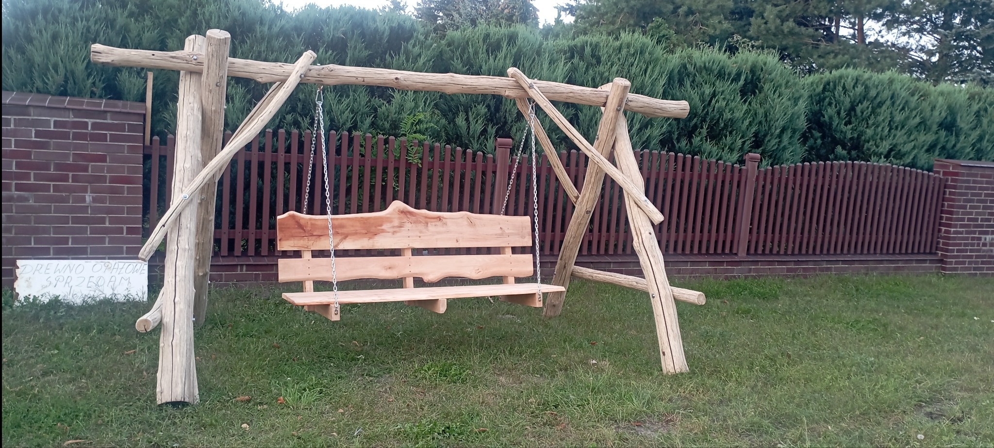 Rustykalna huśtawka ogrodowa z surowego drewna na łańcuchach, na tle zielonego żywopłotu i brązowego płotu. Widoczna tabliczka z napisem.