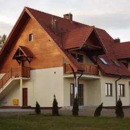 Nowo wybudowany budynek mieszkalny z drewnianą elewacją na poddaszu i czerwonym dachem z dachówki, z balkonami i schodami zewnętrznymi, widok z poziomu gruntu.