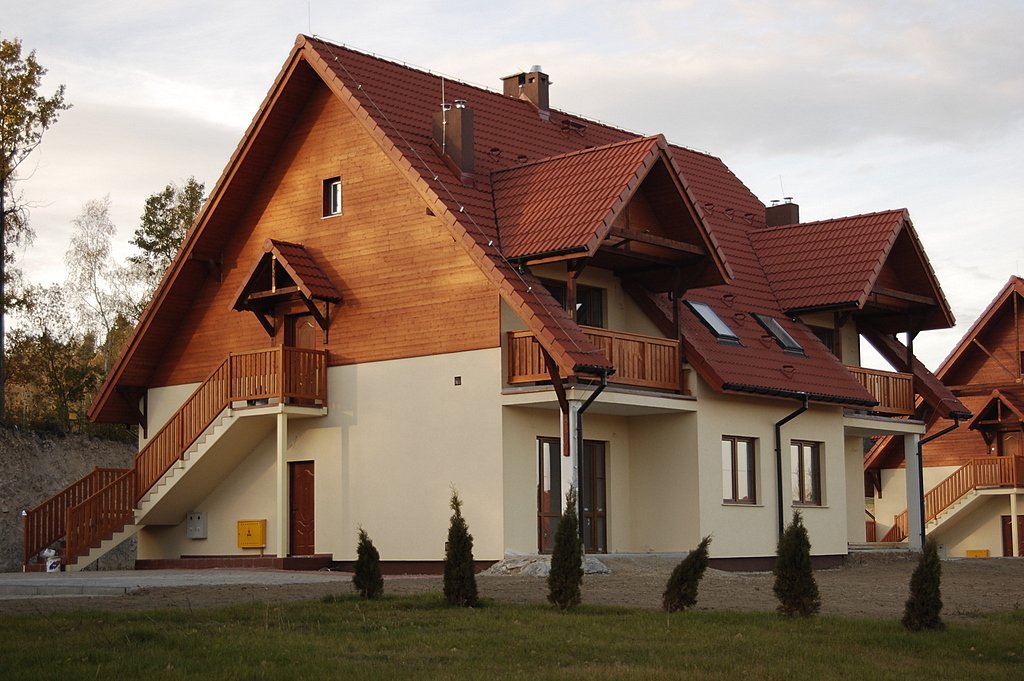 Nowo wybudowany budynek mieszkalny z drewnianą elewacją na poddaszu i czerwonym dachem z dachówki, z balkonami i schodami zewnętrznymi, widok z poziomu gruntu.