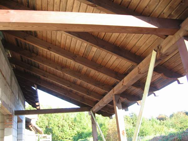 Surowa drewniana konstrukcja dachu z deskowaniem, widok od dołu, częściowo zadaszona przestrzeń, w tle zieleń.