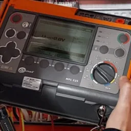 Miernik Sonel MPI-525 w dłoni elektryka, wyświetlający wynik pomiaru 0.0V, na tle panelu sterowniczego z przewodami.