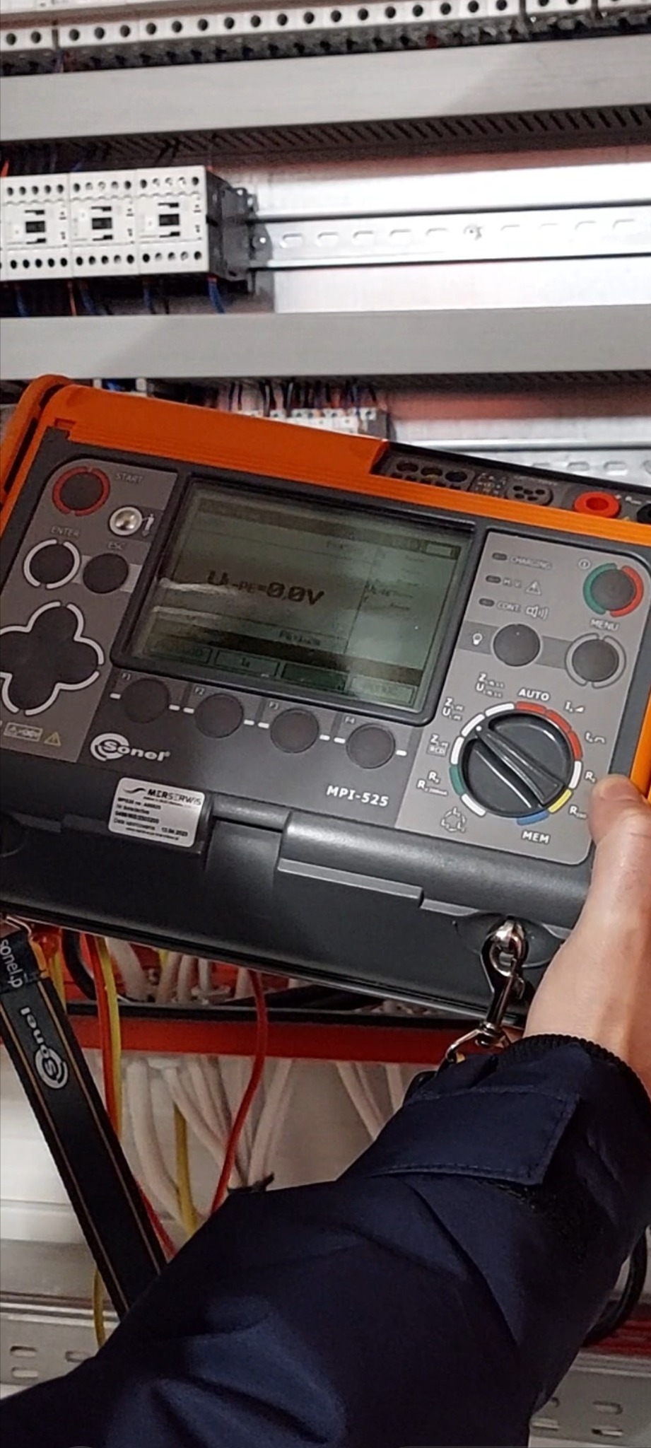 Miernik Sonel MPI-525 w dłoni elektryka, wyświetlający wynik pomiaru 0.0V, na tle panelu sterowniczego z przewodami.