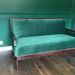 Zakończona renowacja: Sofa dwuosobowa obita zielonym aksamitem, z drewnianymi nogami i ramą, wykończona ozdobnymi ćwiekami, na tle zielonej ściany z boazerią.
