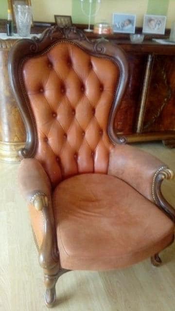 Antyczny fotel z brązowej skóry, pikowany na oparciu, z ozdobnymi drewnianymi elementami, ustawiony w pomieszczeniu z klasycznymi meblami.