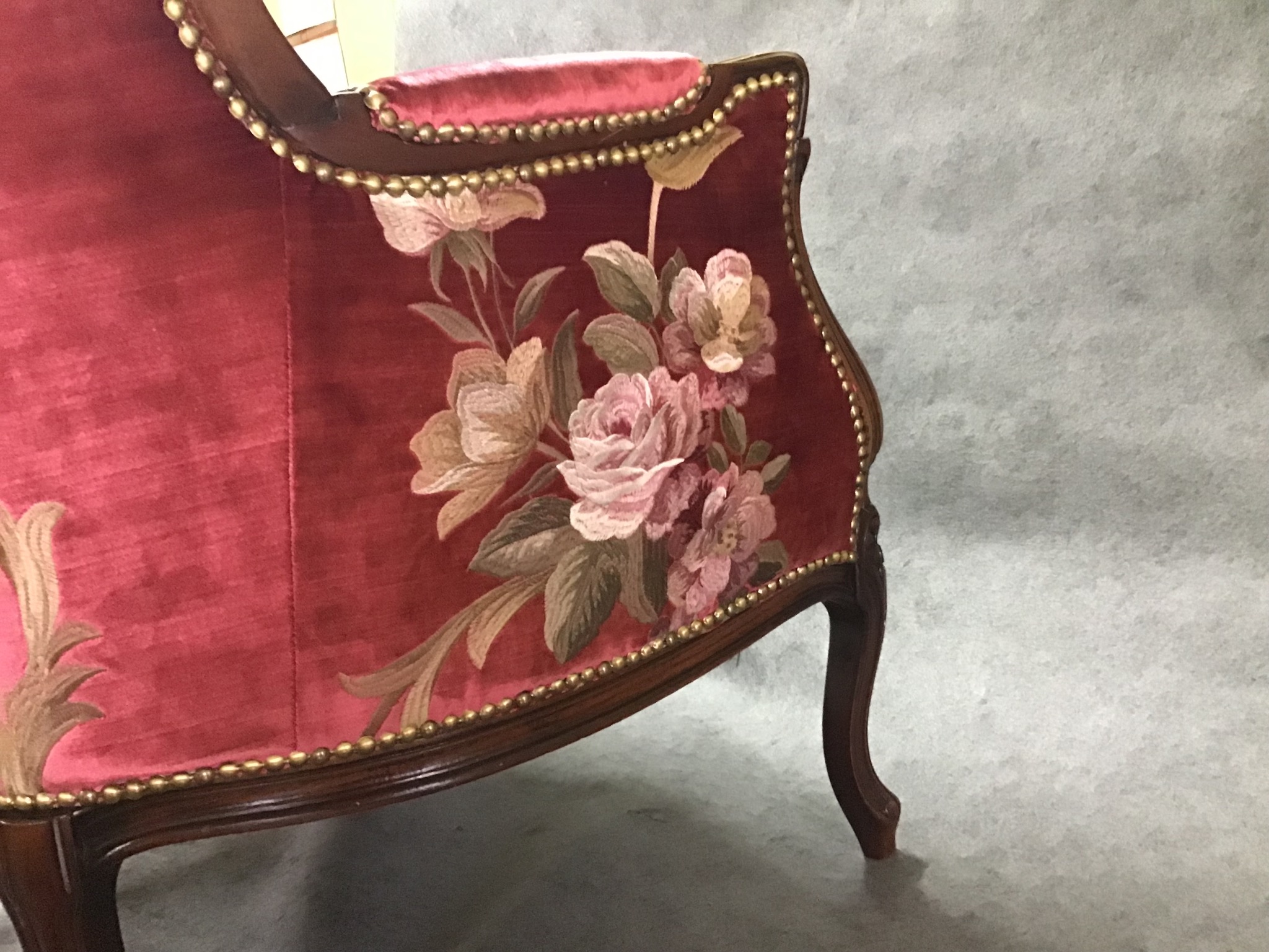 Fragment odrestaurowanego fotela z bordową tapicerką zdobioną haftem z motywem róż i brązowymi gwoździami tapicerskimi wzdłuż krawędzi.