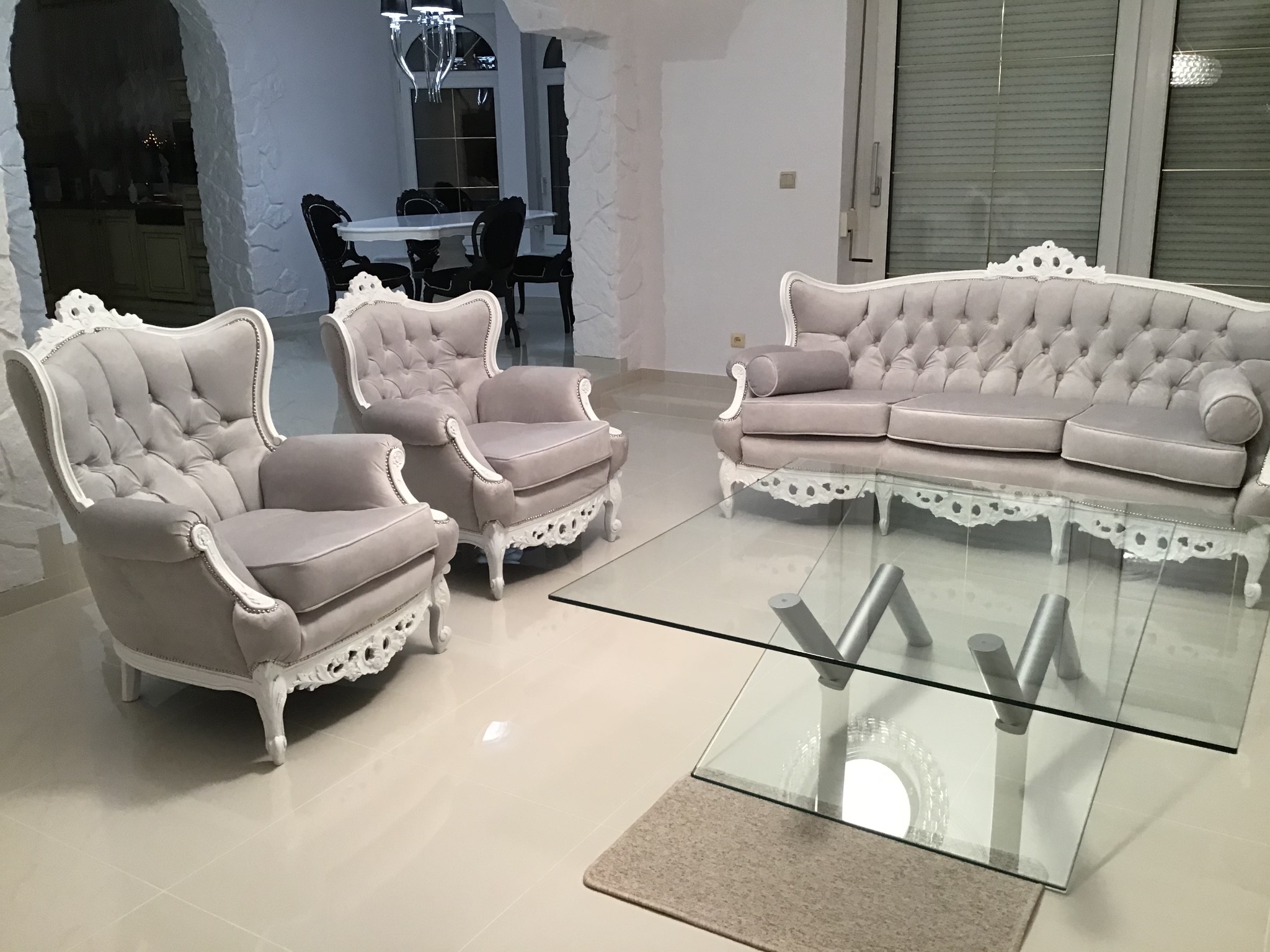Jasnoszary, pikowany komplet wypoczynkowy w stylu Ludwika XVI: sofa i dwa fotele z białymi, rzeźbionymi ramami, ustawione w przestronnym, białym salonie z szklanym stolikiem kawowym na tle jadalni.