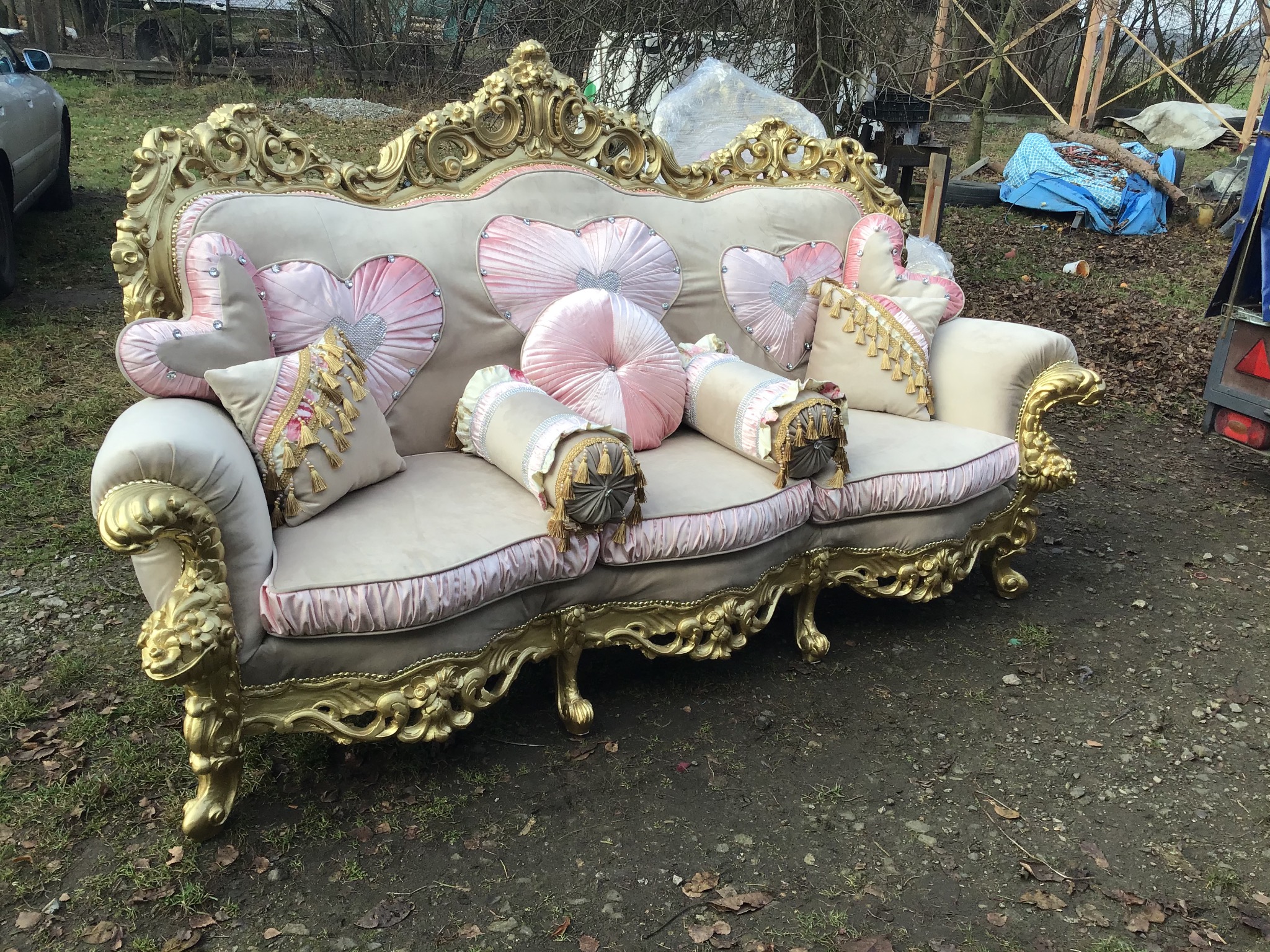 Bogato zdobiona, beżowo-złota sofa w stylu Ludwika, z dekoracyjnymi poduszkami w kształcie serc i wałków, ustawiona na ziemi z liśćmi i gałęziami w tle.