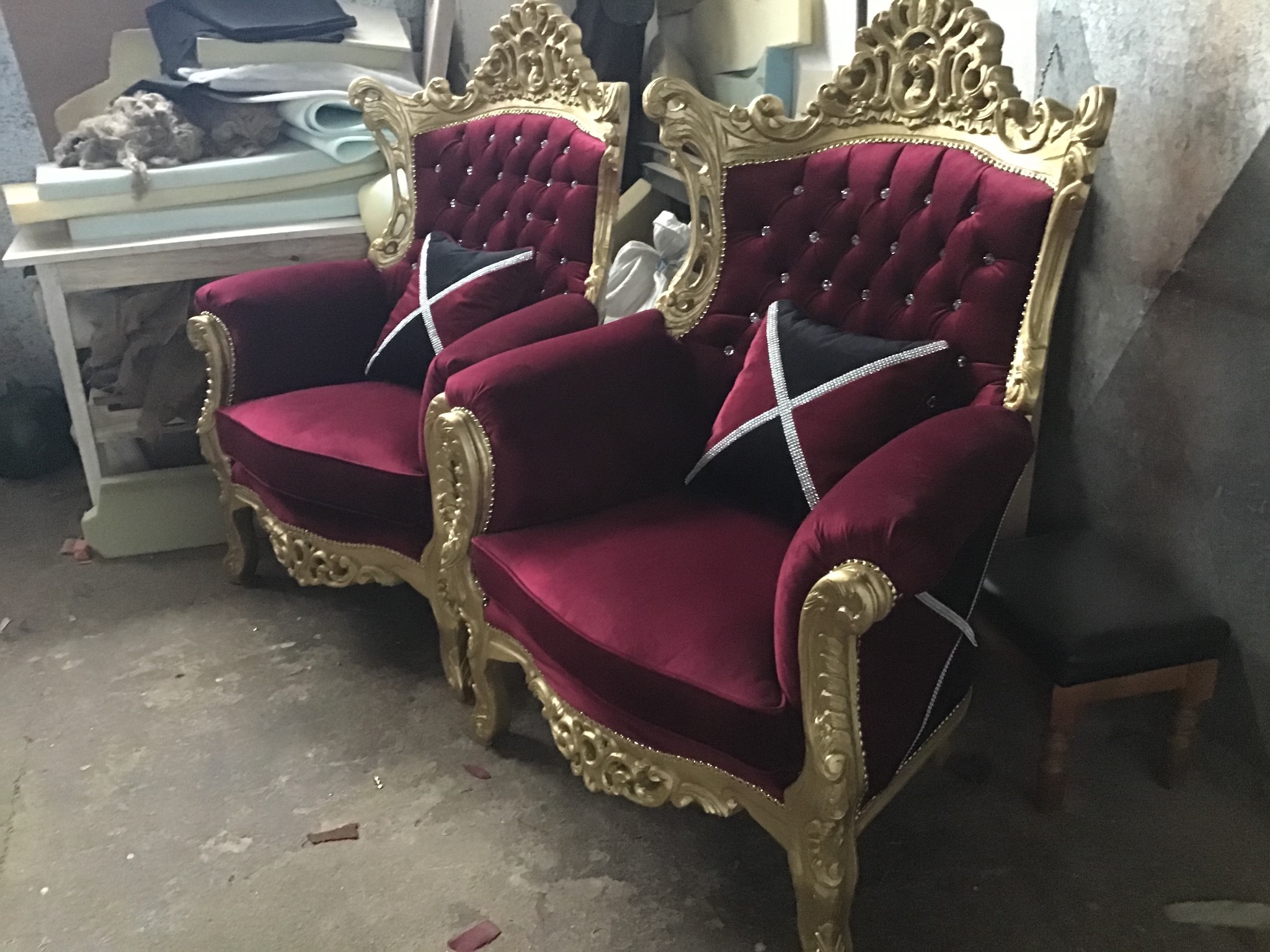 Dwa bordowe fotele w stylu glamour z pikowanym oparciem i złotymi zdobieniami, widoczne poduszki z czarno-białym wzorem, w tle stół roboczy z materiałami tapicerskimi.