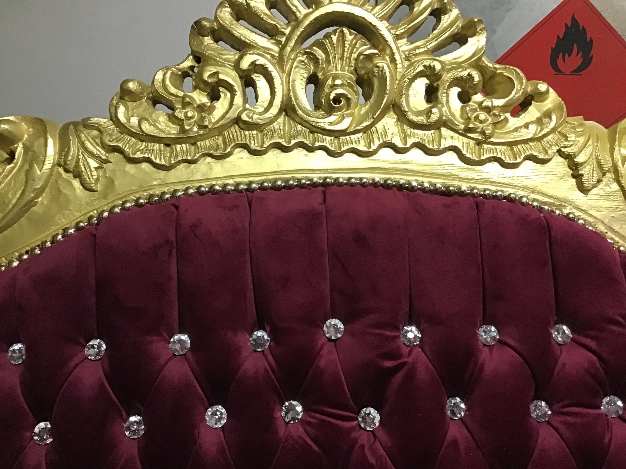Fragment zagłówka fotela w stylu glamour: bordowy welur pikowany kryształkami, złota, zdobiona rama, w tle czerwona tabliczka ostrzegawcza.
