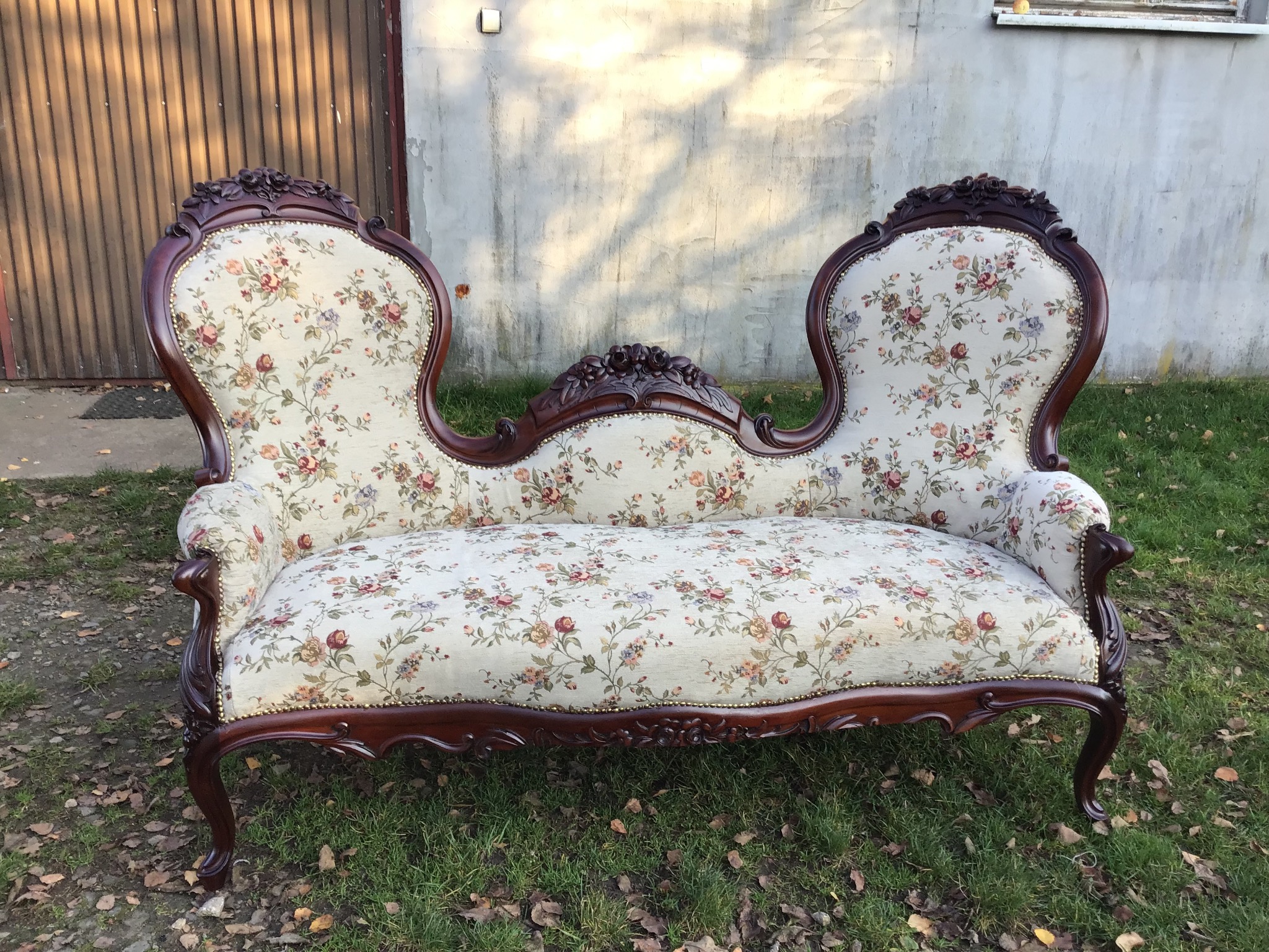 Antyczna kanapa dwuosobowa z ciemnego drewna i tapicerką w kwiaty, stojąca na trawie przed garażem.
