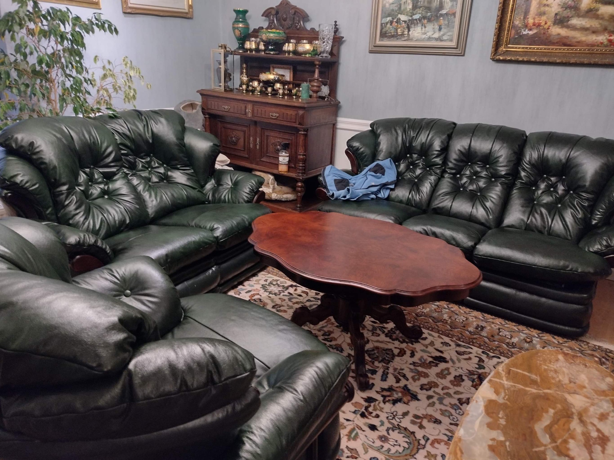 Zestaw wypoczynkowy z ciemnozielonej skóry, składający się z sofy i foteli, ustawiony w pokoju z dywanem w orientalne wzory i antykami w tle.