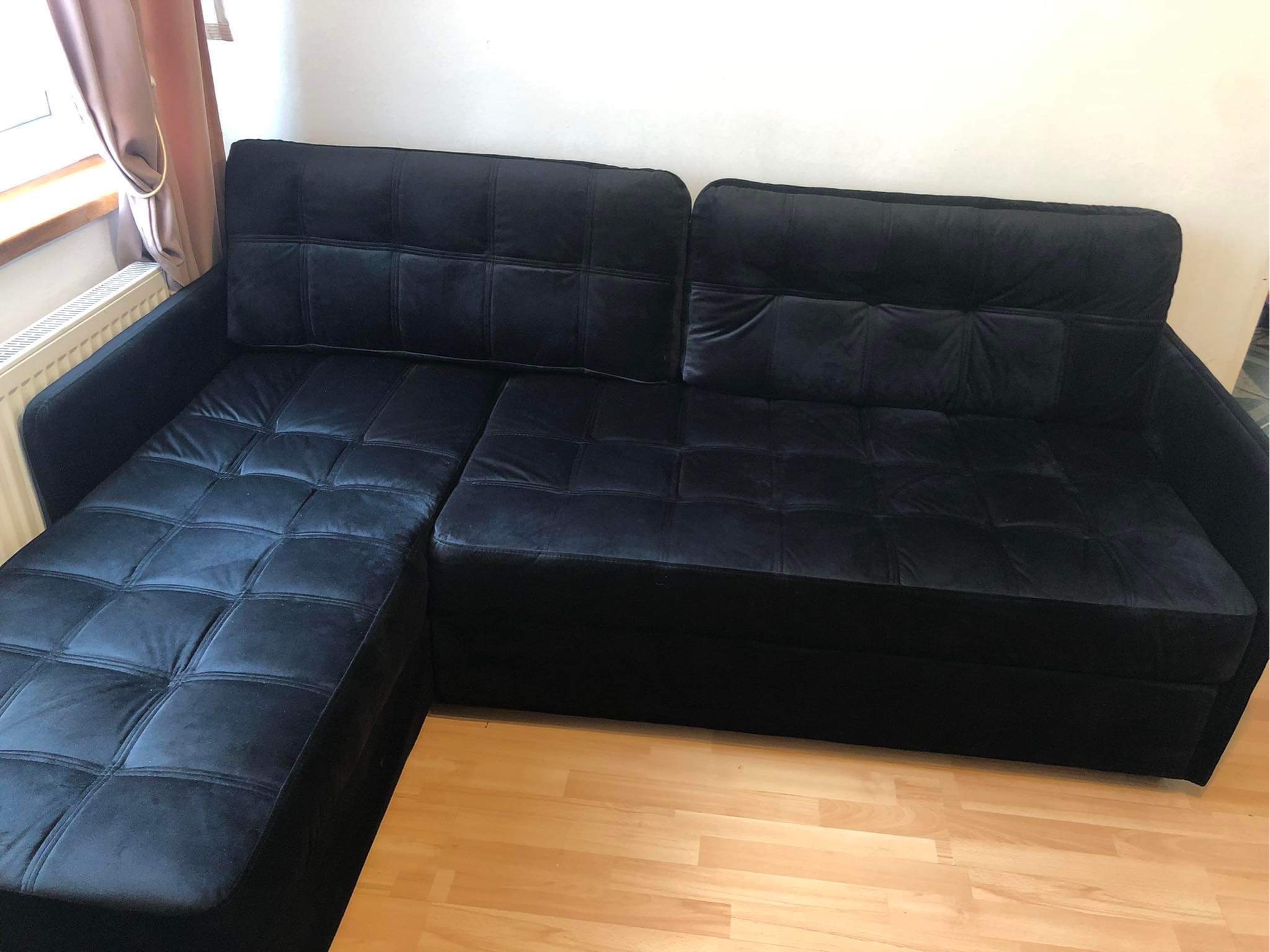 Czarna, welurowa sofa narożna z pikowanymi siedziskami i oparciami, widoczna na drewnianej podłodze w jasnym wnętrzu.