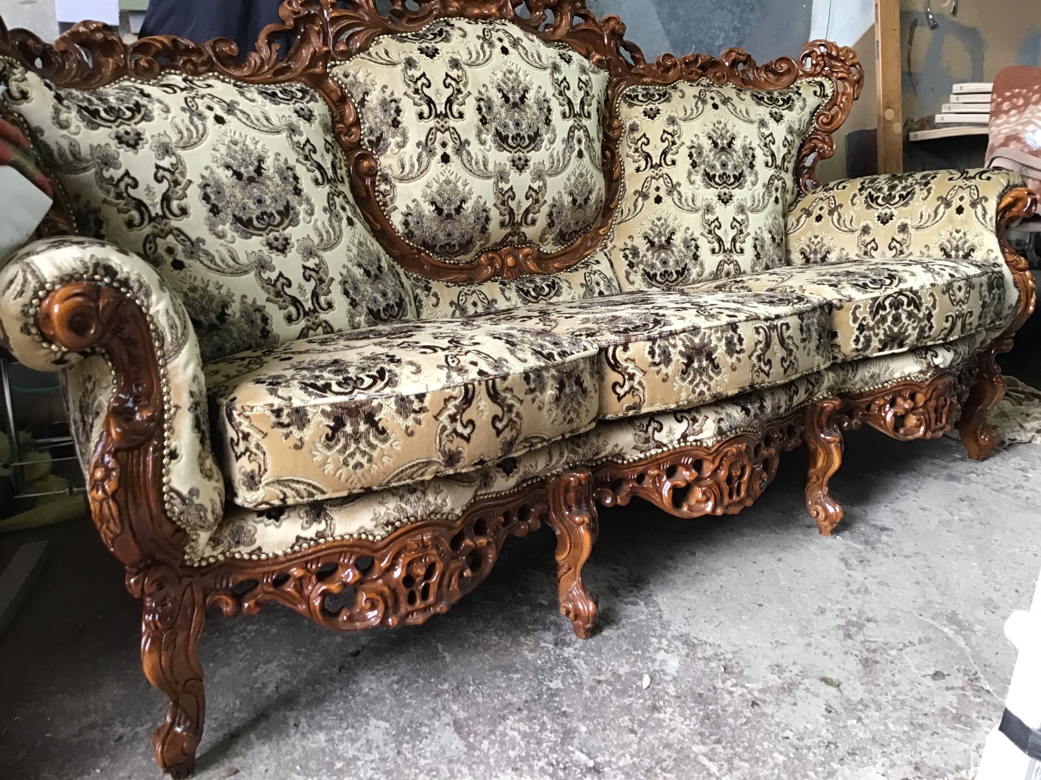 Antyczny, trzysiedziskowy fotel z rzeźbioną, brązową ramą i beżowo-brązową tapicerką w kwiatowe wzory, widoczny z boku, na szarym, betonowym tle.