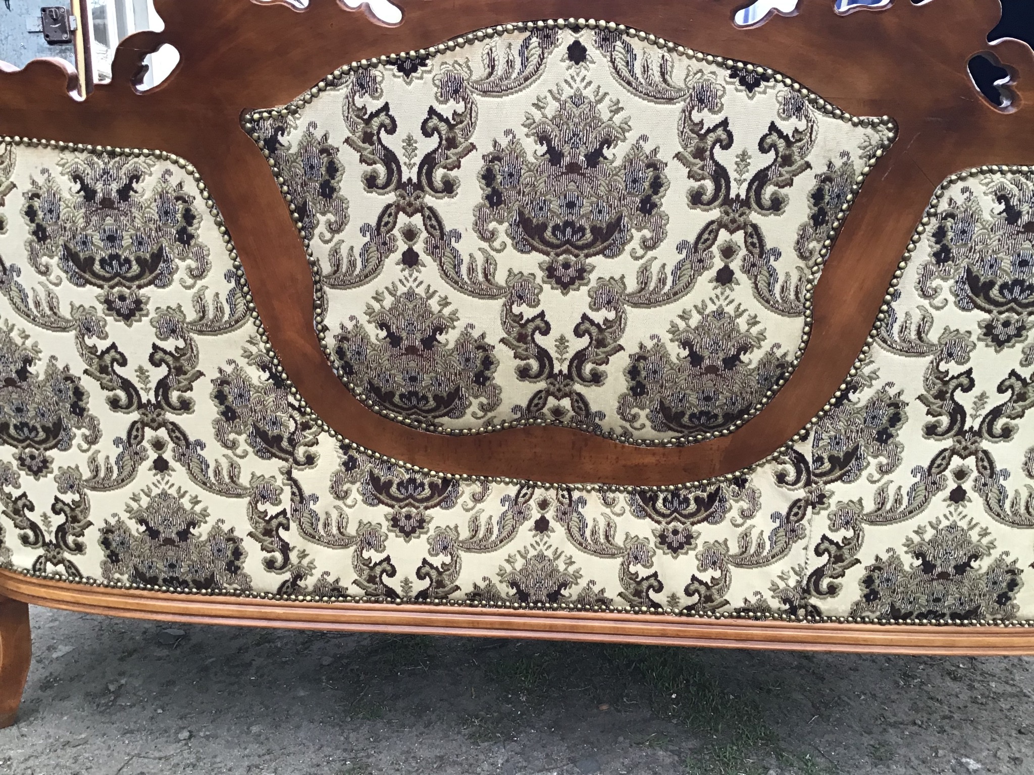 Fragment odnowionego fotela z drewnianą ramą i wzorzystą, kremowo-brązową tapicerką, wykończoną ozdobnymi ćwiekami.