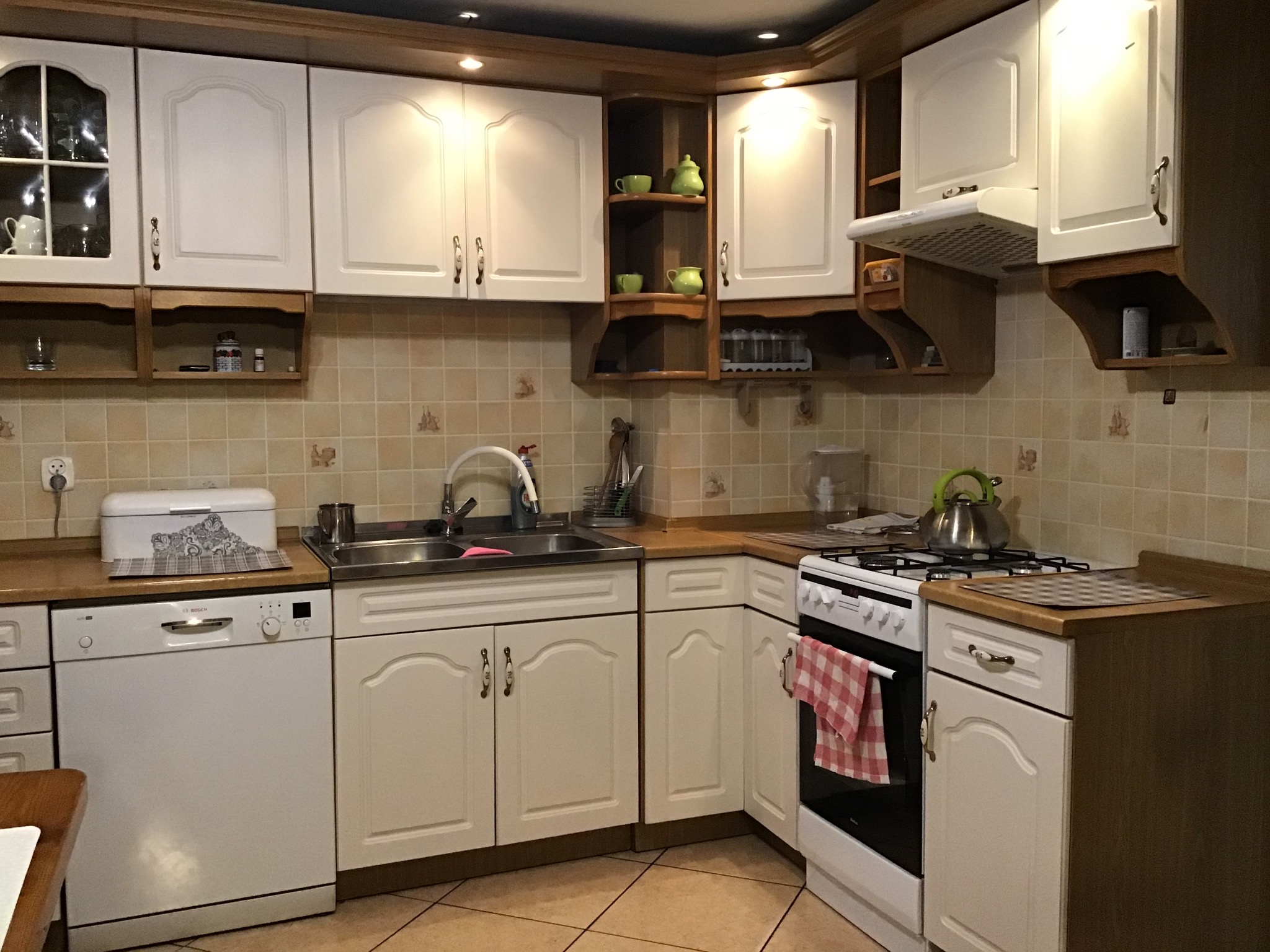 Kuchnia z białymi, frezowanymi frontami szafek, brązowymi blatami i beżowymi płytkami na ścianach, widok na zlewozmywak, kuchenkę i zmywarkę.
