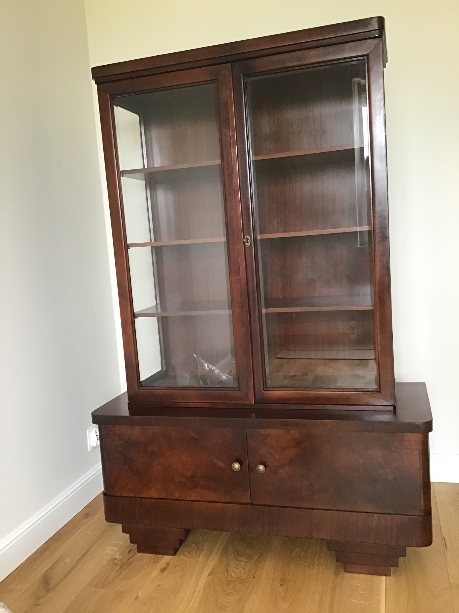 Antyczna, dwuczęściowa witryna z ciemnego drewna z przeszkloną górną częścią i dolną zamykaną na dwa mosiężne uchwyty, ustawiona na drewnianej podłodze przy jasnej ścianie.