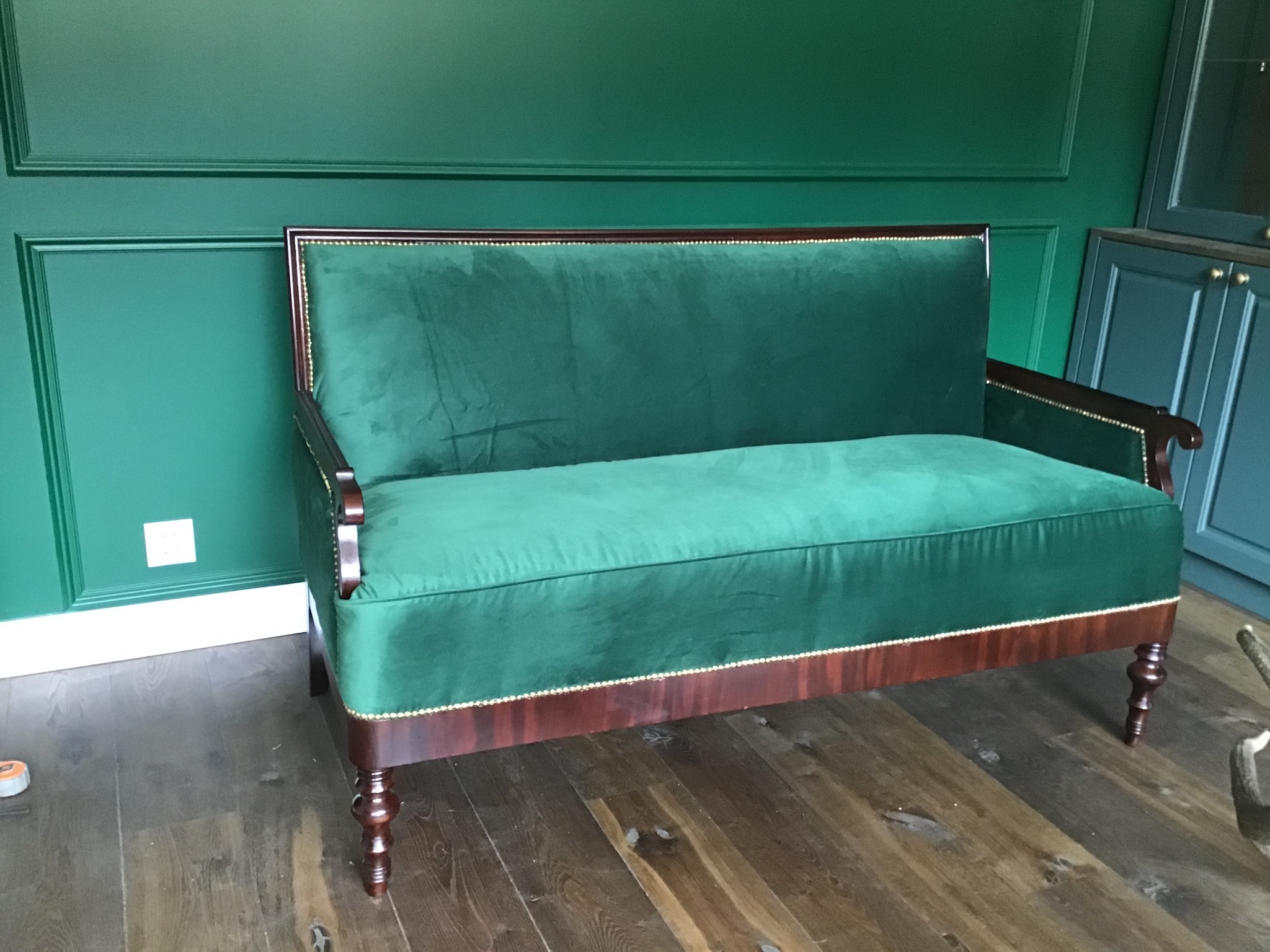 Zakończona renowacja: Sofa dwuosobowa obita zielonym aksamitem, z drewnianymi nogami i ramą, wykończona ozdobnymi ćwiekami, na tle zielonej ściany z boazerią.
