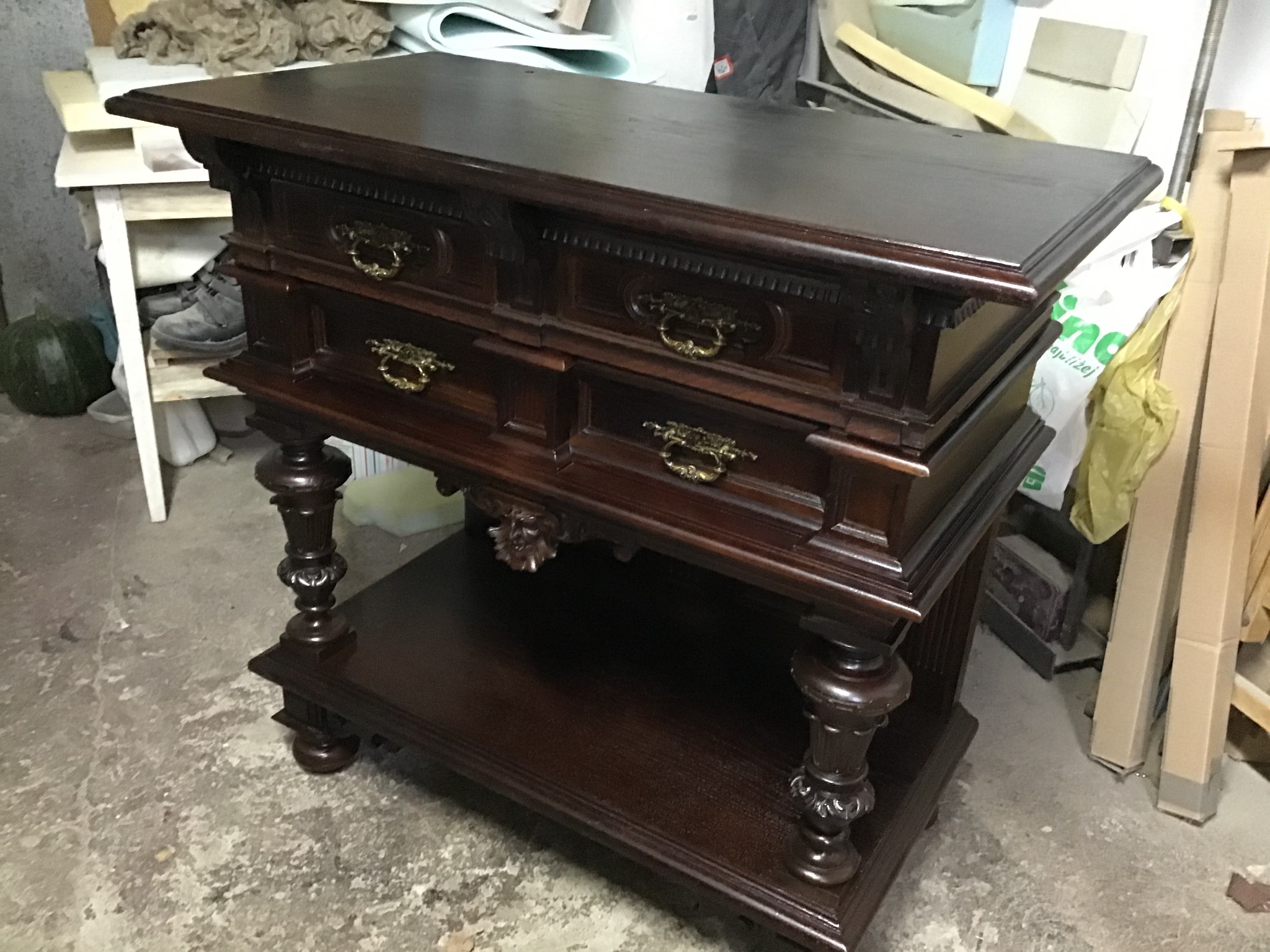 Odrestaurowana, ciemnobrązowa komoda z szufladami i rzeźbionymi nogami, widoczna w warsztacie w Galewicach, z elementami tapicerskimi w tle.