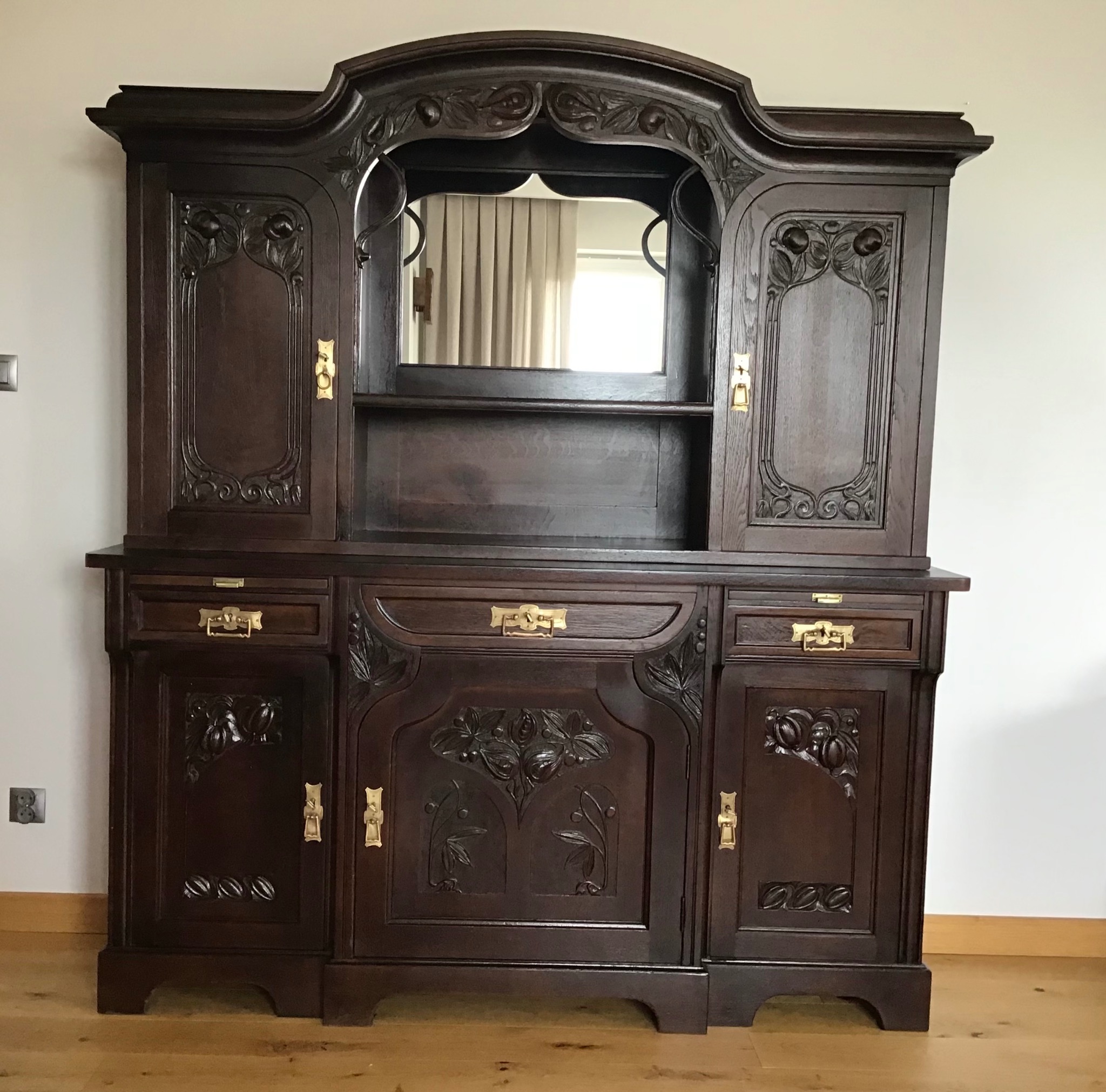 Ciemnobrązowy, rzeźbiony kredens w stylu art deco z lustrem, złotymi uchwytami i misternymi zdobieniami kwiatowymi na frontach szafek.