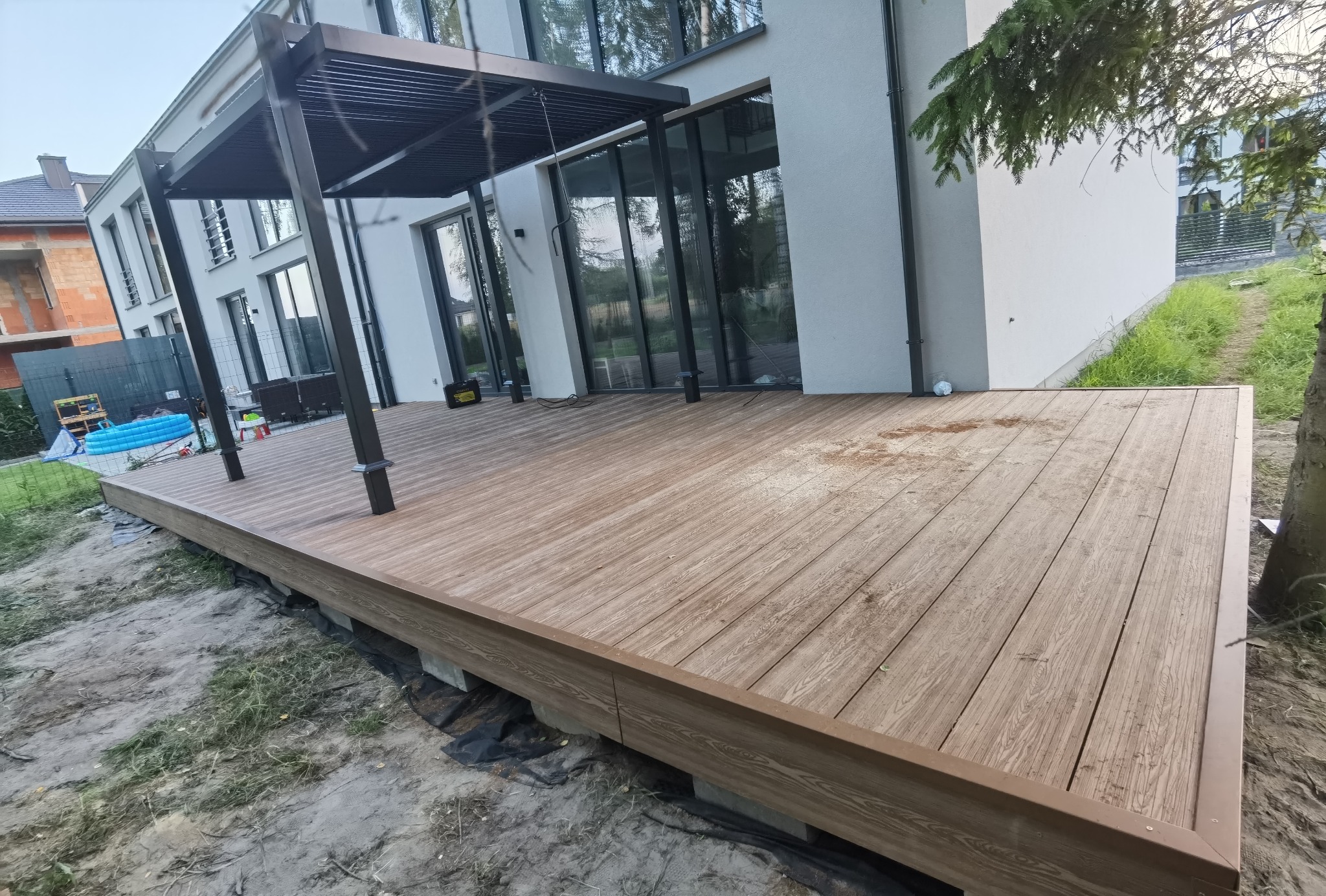 Nowoczesny taras kompozytowy z pergolą przy białym domu z dużymi oknami, widoczne niedokończone prace ziemne wokół tarasu.
