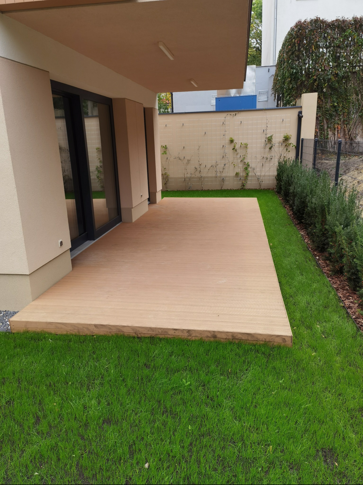 Nowoczesny taras z jasnego drewna przylegający do budynku, otoczony świeżo skoszoną trawą i niskimi krzewami.