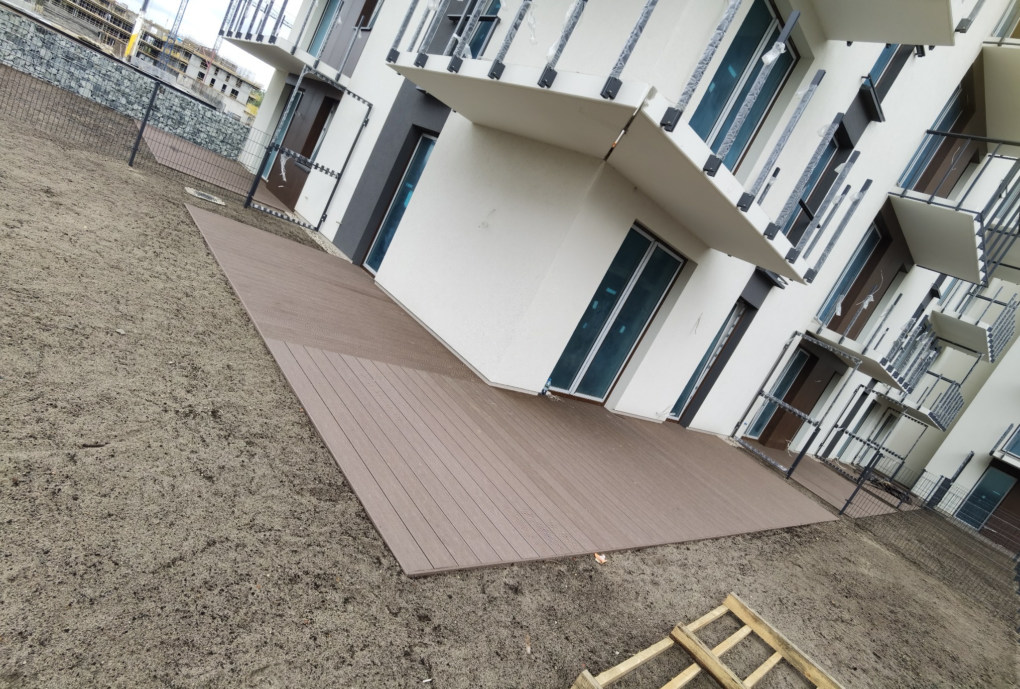 Nowoczesny taras z desek kompozytowych przylegający do budynku wielorodzinnego, widok z poziomu gruntu na niezagospodarowany teren.