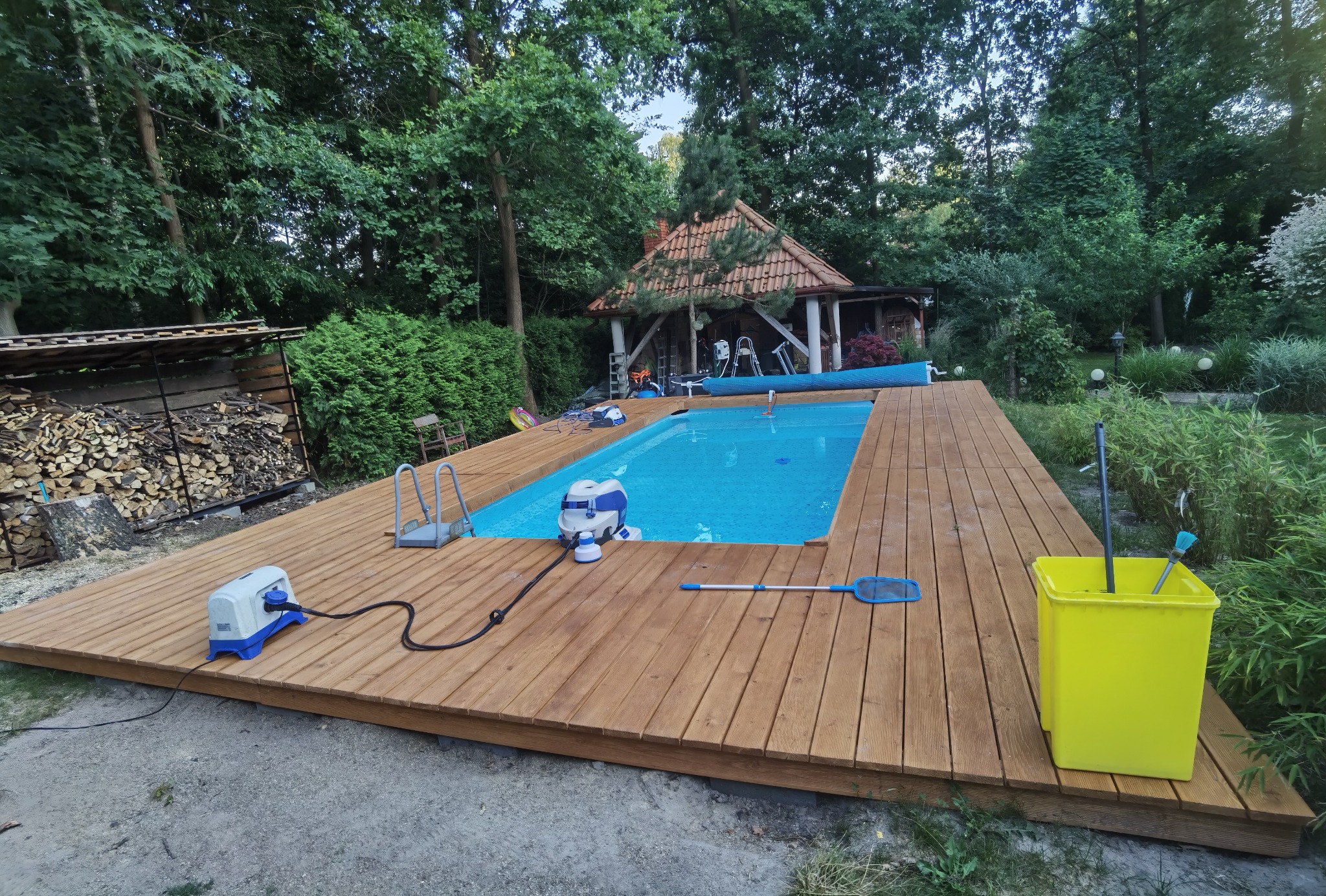 Basen otoczony nowym drewnianym tarasem w ogrodzie, widoczne akcesoria do czyszczenia basenu i drewutnia w tle.
