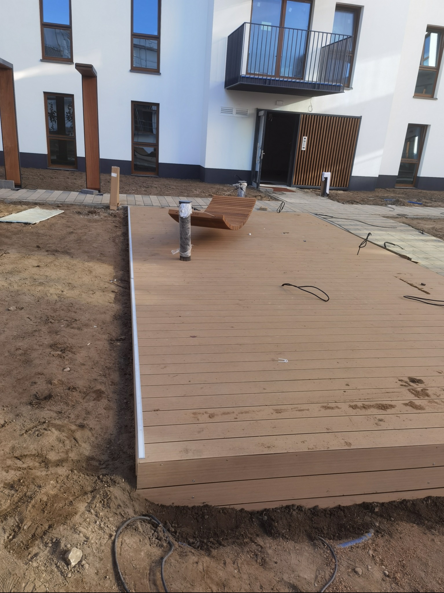 Montaż tarasu kompozytowego przy nowoczesnym budynku mieszkalnym, widoczne kable i rury w ziemi, ławka z tego samego materiału, etap prac wykończeniowych.