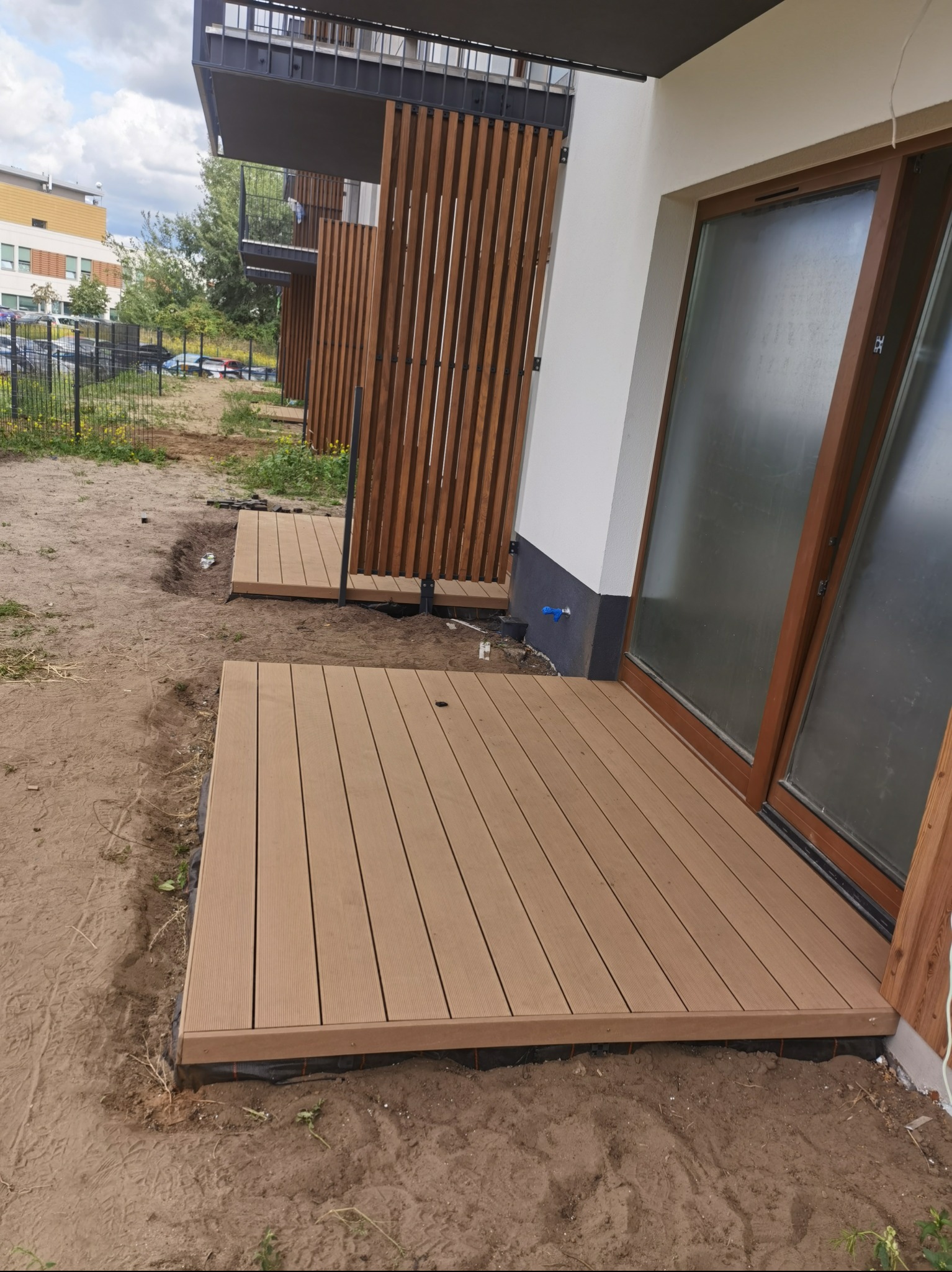 Montaż tarasu kompozytowego przy nowoczesnym budynku mieszkalnym, widoczne deski tarasowe w kolorze jasnego drewna, czarna mata geowłókninowa pod konstrukcją, przygotowany teren wokół tarasu.