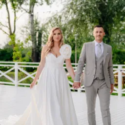 Para młoda trzymająca się za ręce spaceruje po białym tarasie w Ząbkach. Panna młoda w białej sukni z ozdobnym kwiatem na ramieniu, pan młody w szarym garniturze.