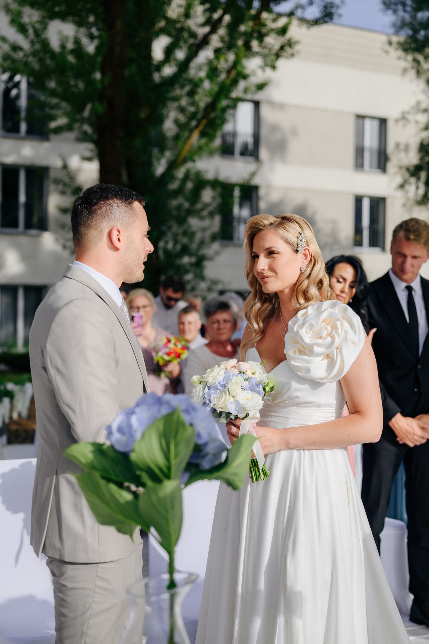Para młoda podczas ceremonii ślubnej na tle budynku z drzewami, panna młoda w białej sukni z ozdobną różą na ramieniu i bukietem, pan młody w beżowym garniturze.