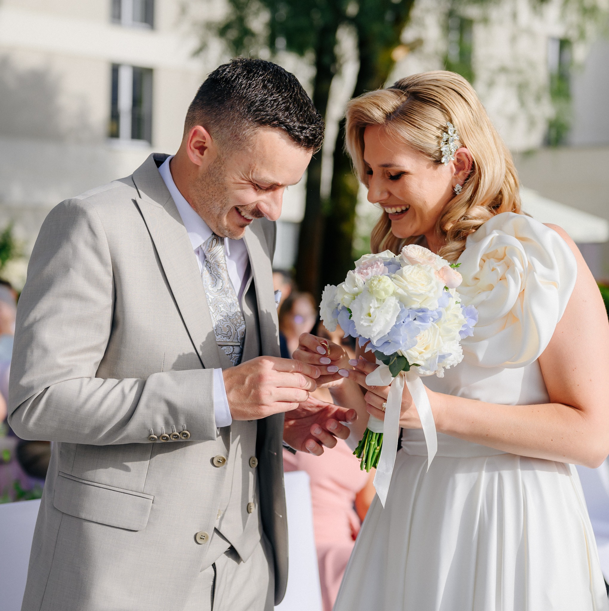 Uśmiechnięta para młoda podczas ceremonii wymiany obrączek, panna młoda w białej sukni z bukietem, pan młody w jasnym garniturze.