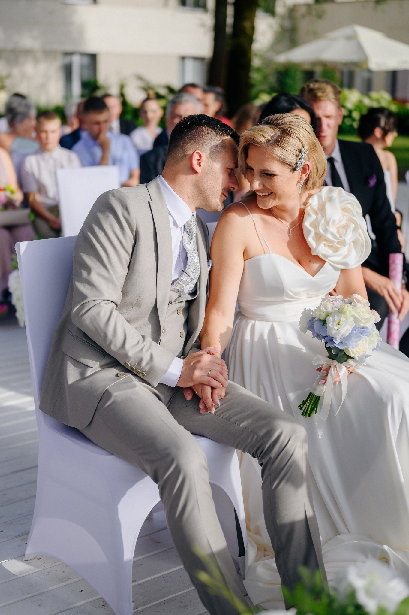 Para młoda siedzi na krzesłach podczas ceremonii ślubnej, trzymając się za ręce, panna młoda z kwiatami i ozdobą we włosach, pan młody w szarym garniturze.
