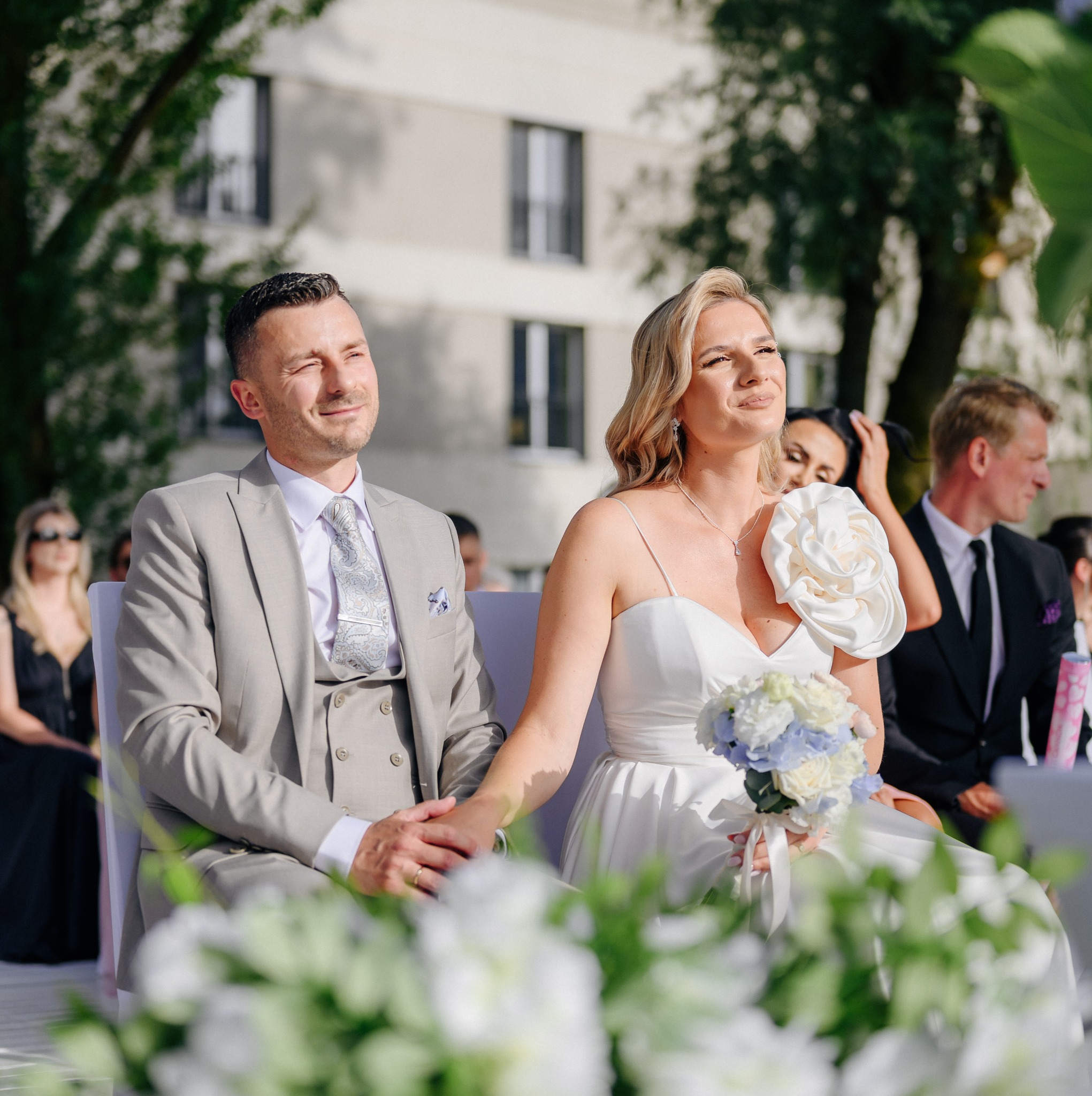 Para młoda siedzi podczas ceremonii ślubnej na tle budynku, trzymając się za ręce, panna młoda ma bukiet z białych i niebieskich kwiatów, a na sukni duży kwiat z materiału.