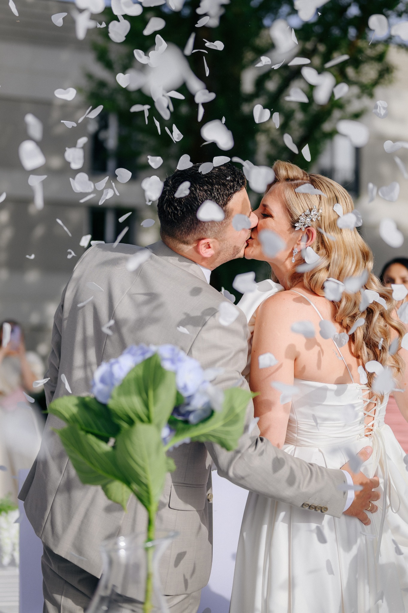Pocałunek pary młodej w deszczu białych konfetti w kształcie serc, panna młoda z upiętymi blond włosami i ozdobną spinką, pan młody w szarym garniturze, bukiet hortensji w tle.
