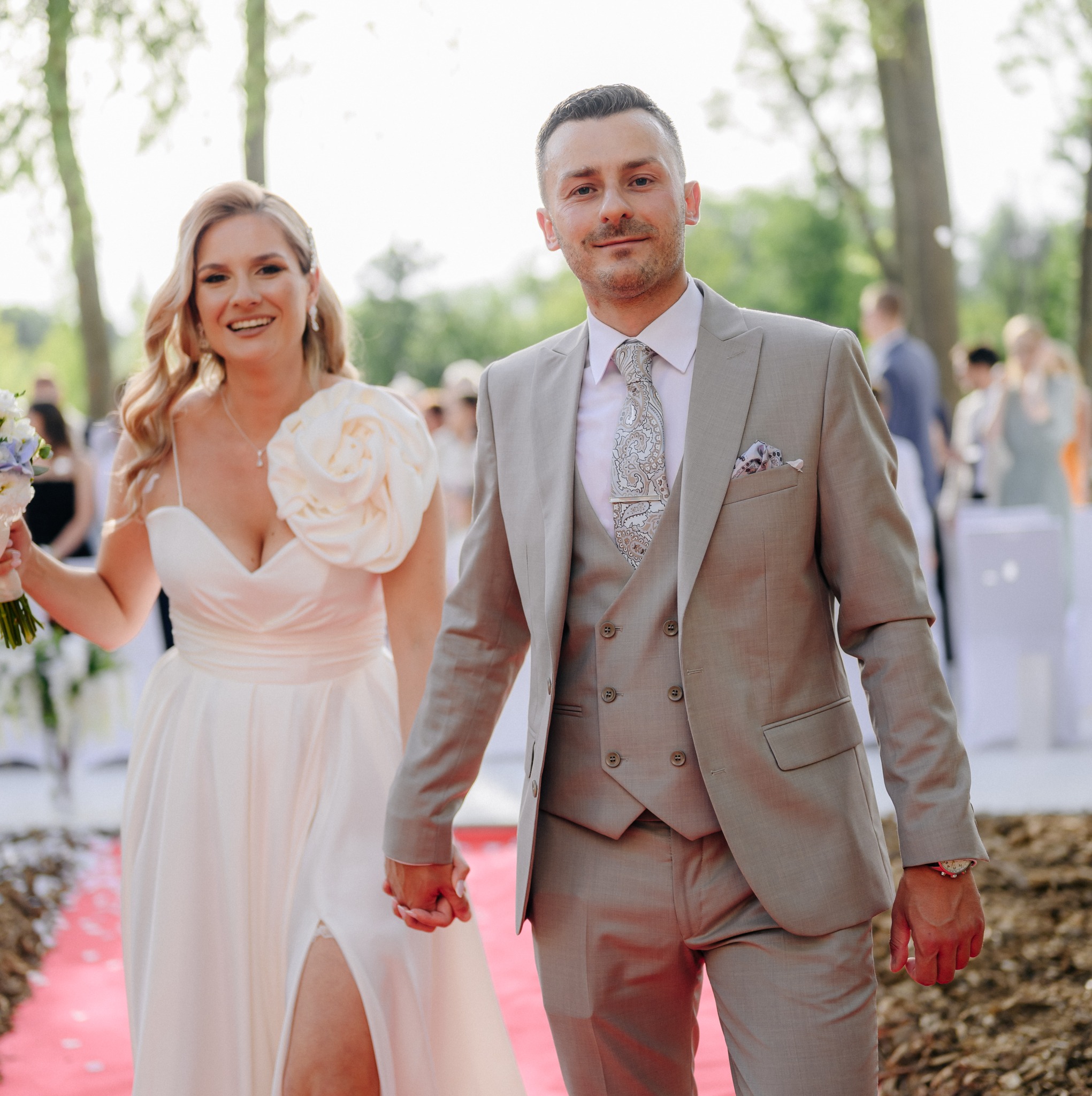 Uśmiechnięta para młoda, on w szarym garniturze z wzorzystym krawatem, ona w białej sukni z kwiatem na ramieniu, trzymają się za ręce na czerwonym dywanie podczas ceremonii ślubnej na świeżym...