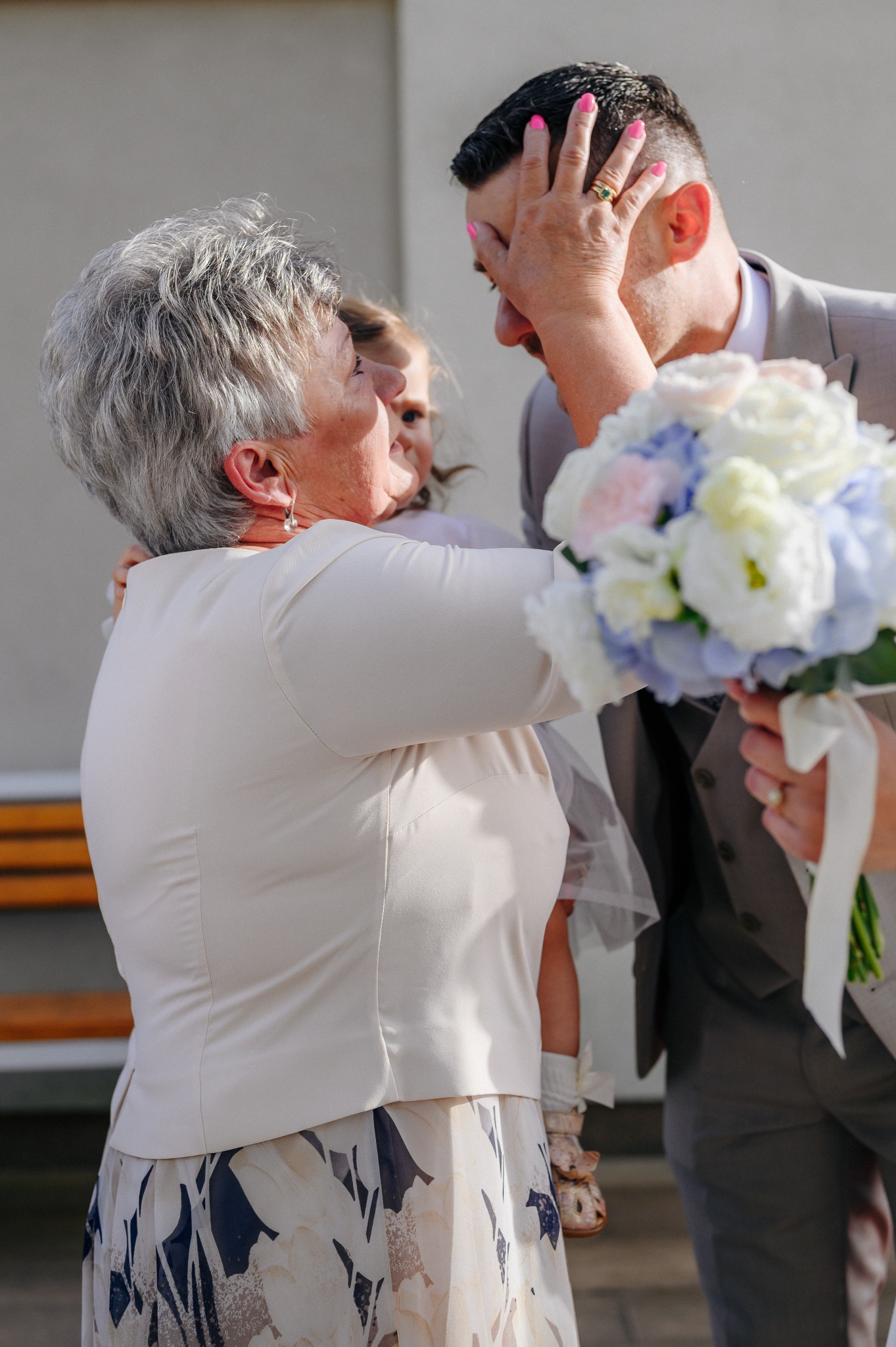 Wzruszający moment po ceremonii ślubnej: panna młoda w jasnej sukni, trzyma bukiet białych i niebieskich kwiatów, obejmowana przez wzruszoną kobietę z krótkimi siwymi włosami, obok stoi mała...