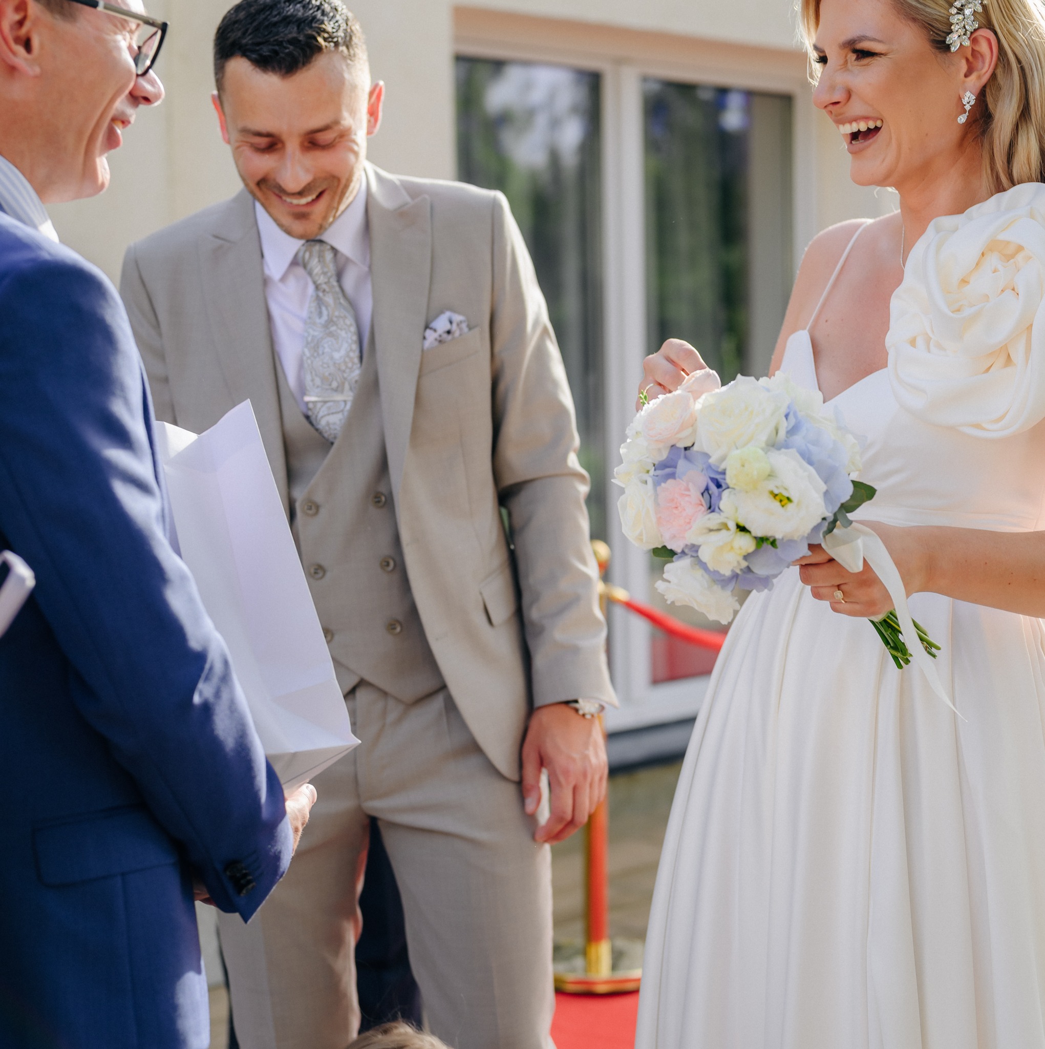 Uśmiechnięta panna młoda w białej sukni z kwiatami, obok pan młody w beżowym garniturze i mężczyzna w niebieskiej marynarce z białą torbą, wszyscy radośnie spoglądają w dół.