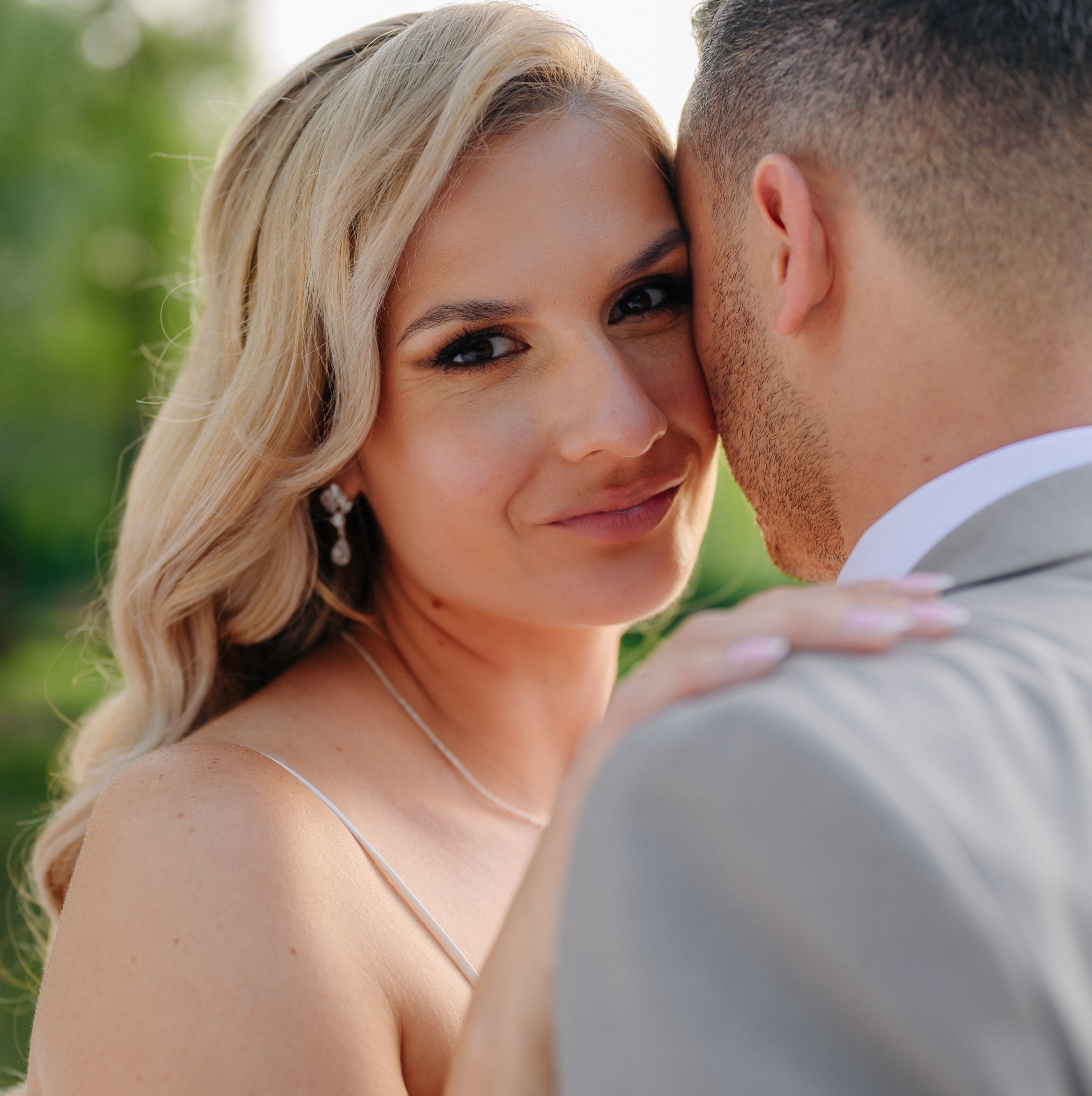 Portret uśmiechniętej blondynki w białej sukience, opierającej głowę o ramię mężczyzny w szarym garniturze, z zielonym tłem.