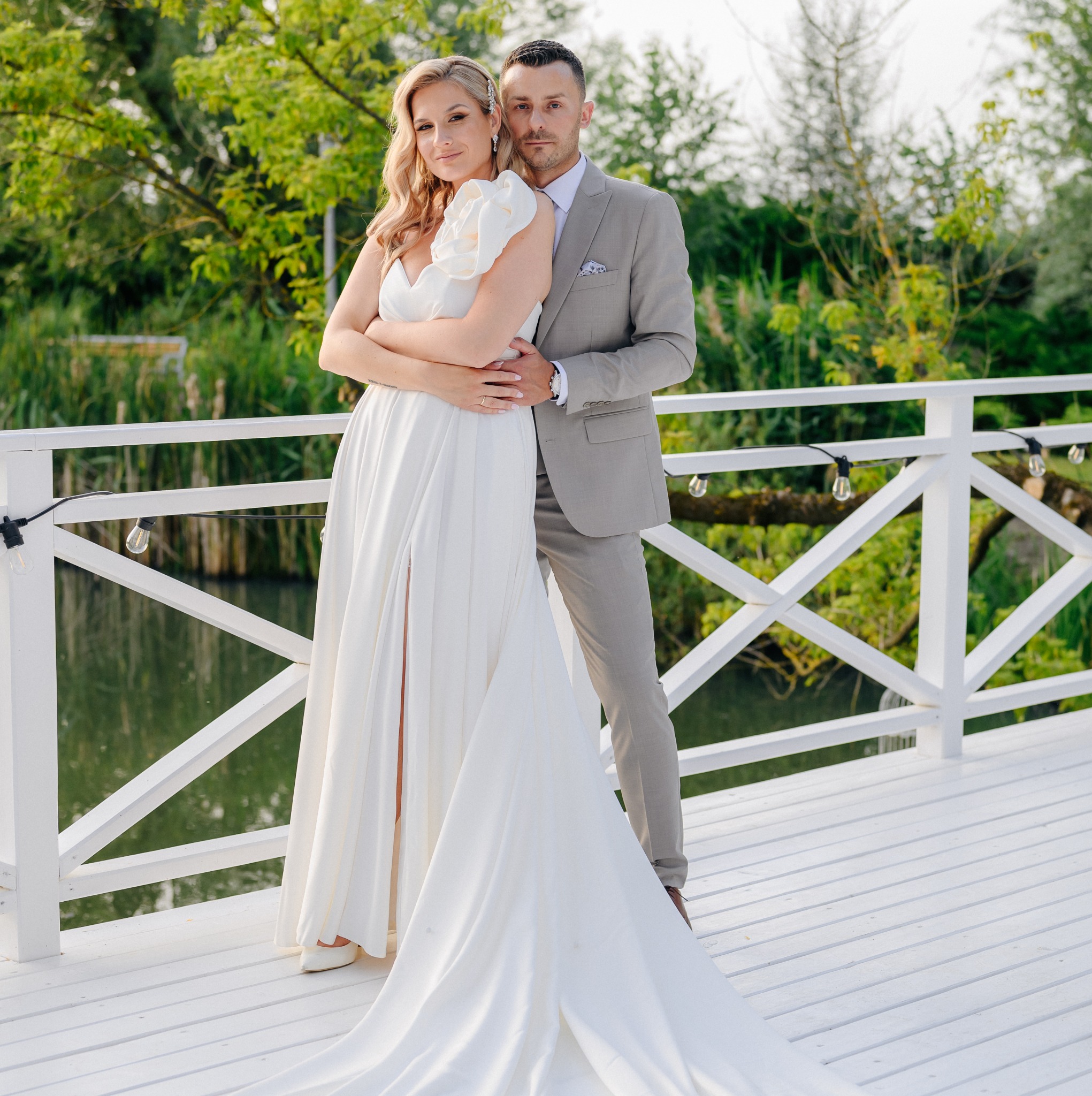 Elegancka para młoda w objęciach na białym pomoście z widokiem na wodę i zieleń, panna młoda w sukni z rozcięciem i ozdobnym kwiatem na ramieniu, pan młody w szarym garniturze.