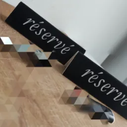 Dwa czarne, drewniane oznaczenia 'réservé' z białą, ozdobną czcionką, ustawione na drewnianym blacie. Widoczne delikatne refleksy świetlne.