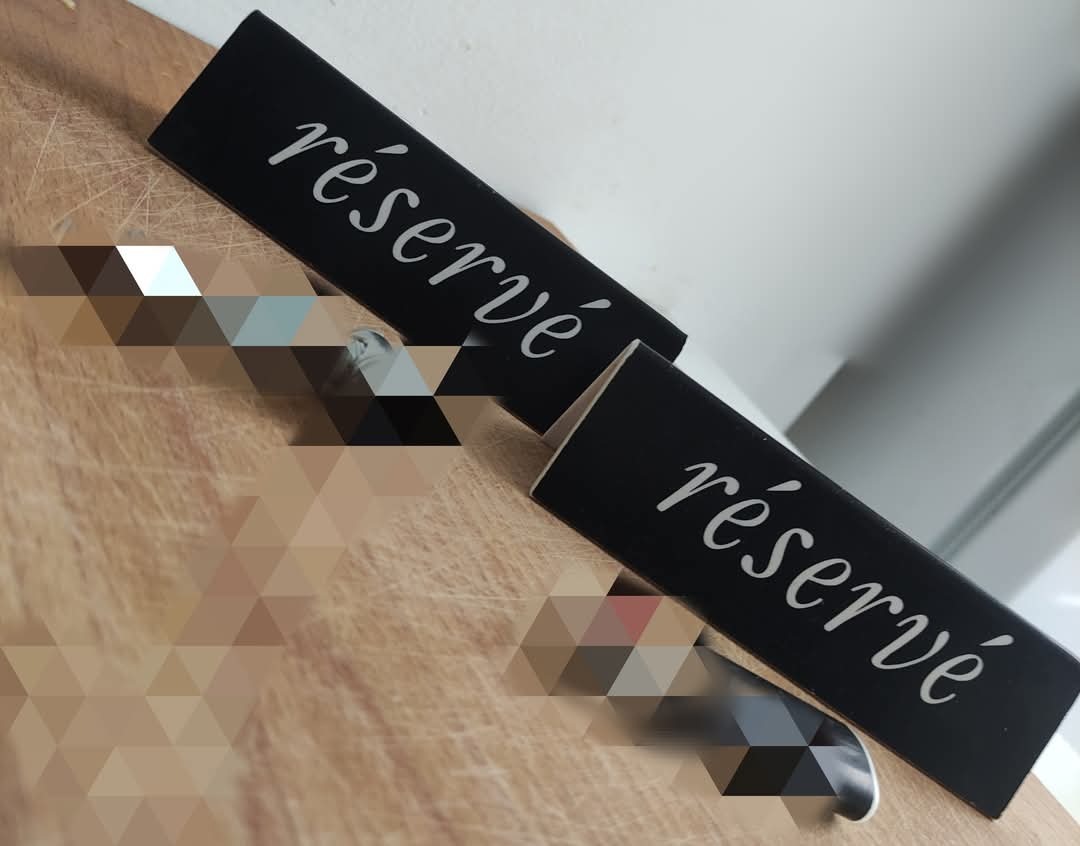 Dwa czarne, drewniane oznaczenia 'réservé' z białą, ozdobną czcionką, ustawione na drewnianym blacie. Widoczne delikatne refleksy świetlne.