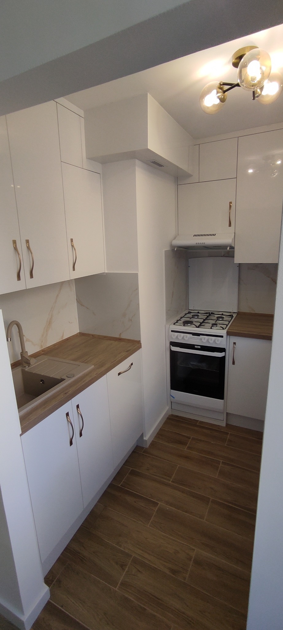 Biała kuchnia z drewnianymi blatami i podłogą. Widoczny zlew, kuchenka i oświetlenie sufitowe. Minimalistyczny design, jasne wnętrze.